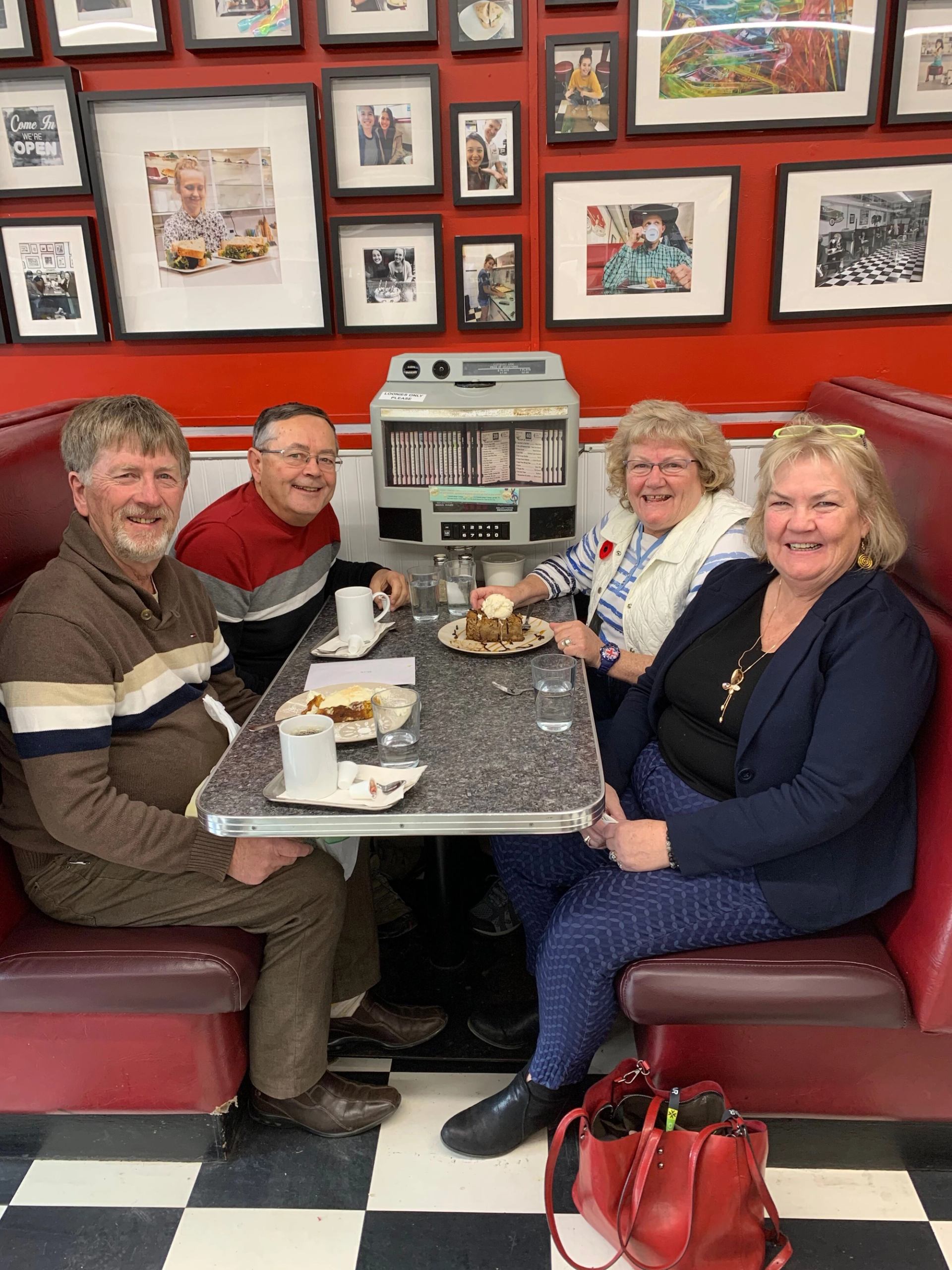 Four adults sit in a red diner booth sharing dessert, with jukebox and photo wall behind.
