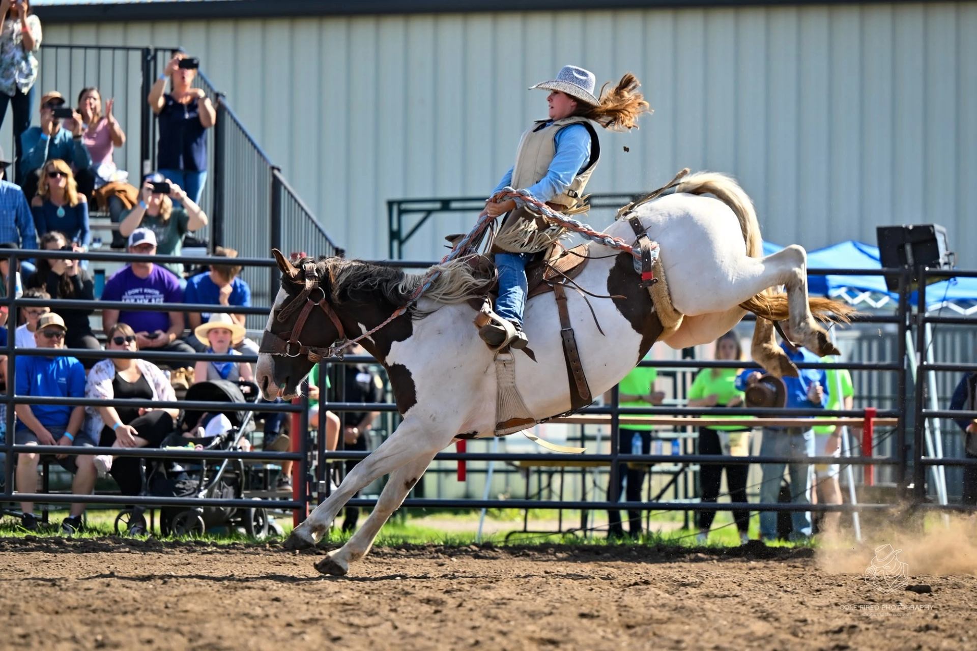 Rodeo rider leans back on bucking horse in dirt arena before metal bleachers.