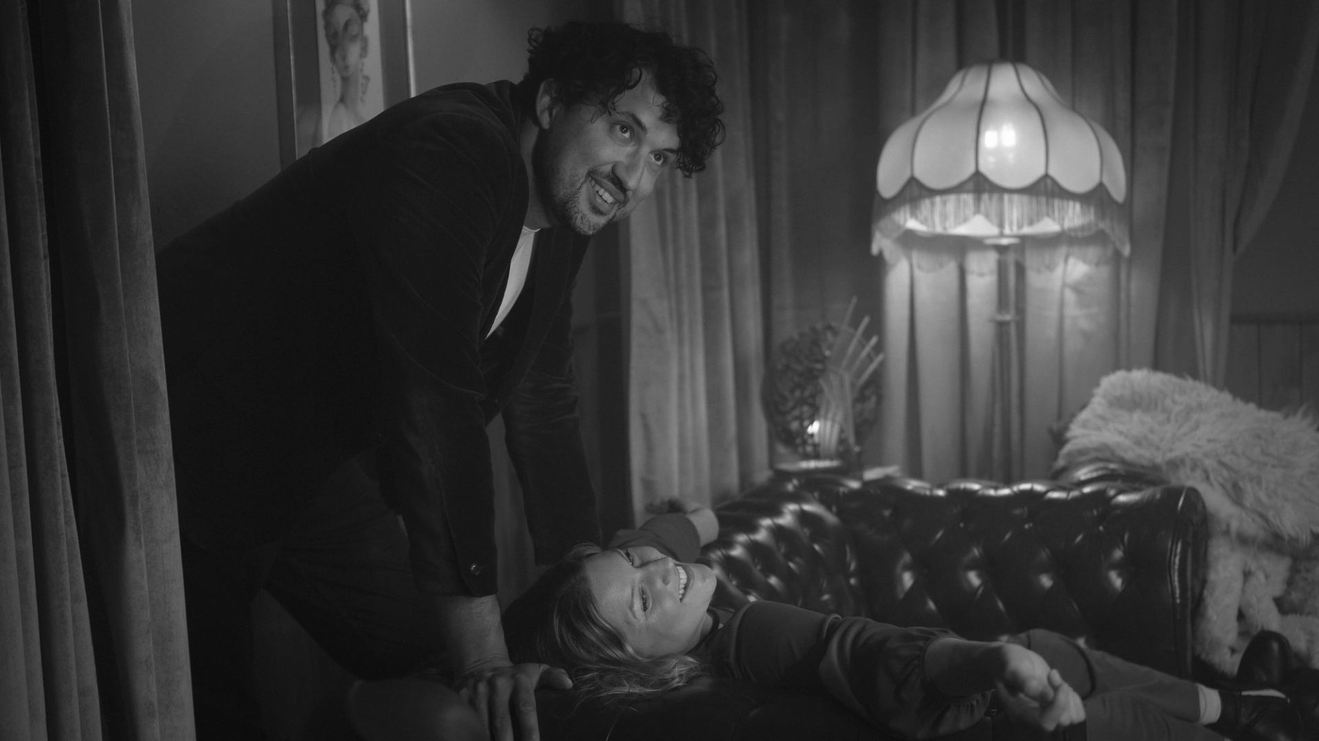 Black-and-white scene of two people beside a leather sofa and glowing floor lamp.