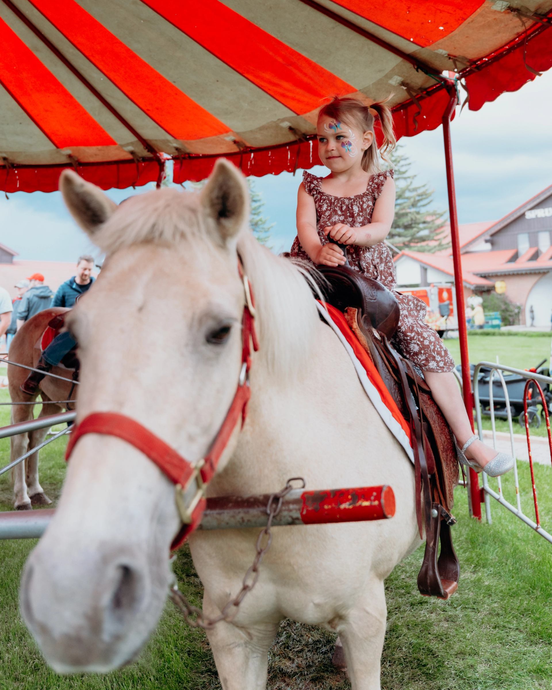 Little girl with face paint rides a white pony under a red-and-white tent at the event.