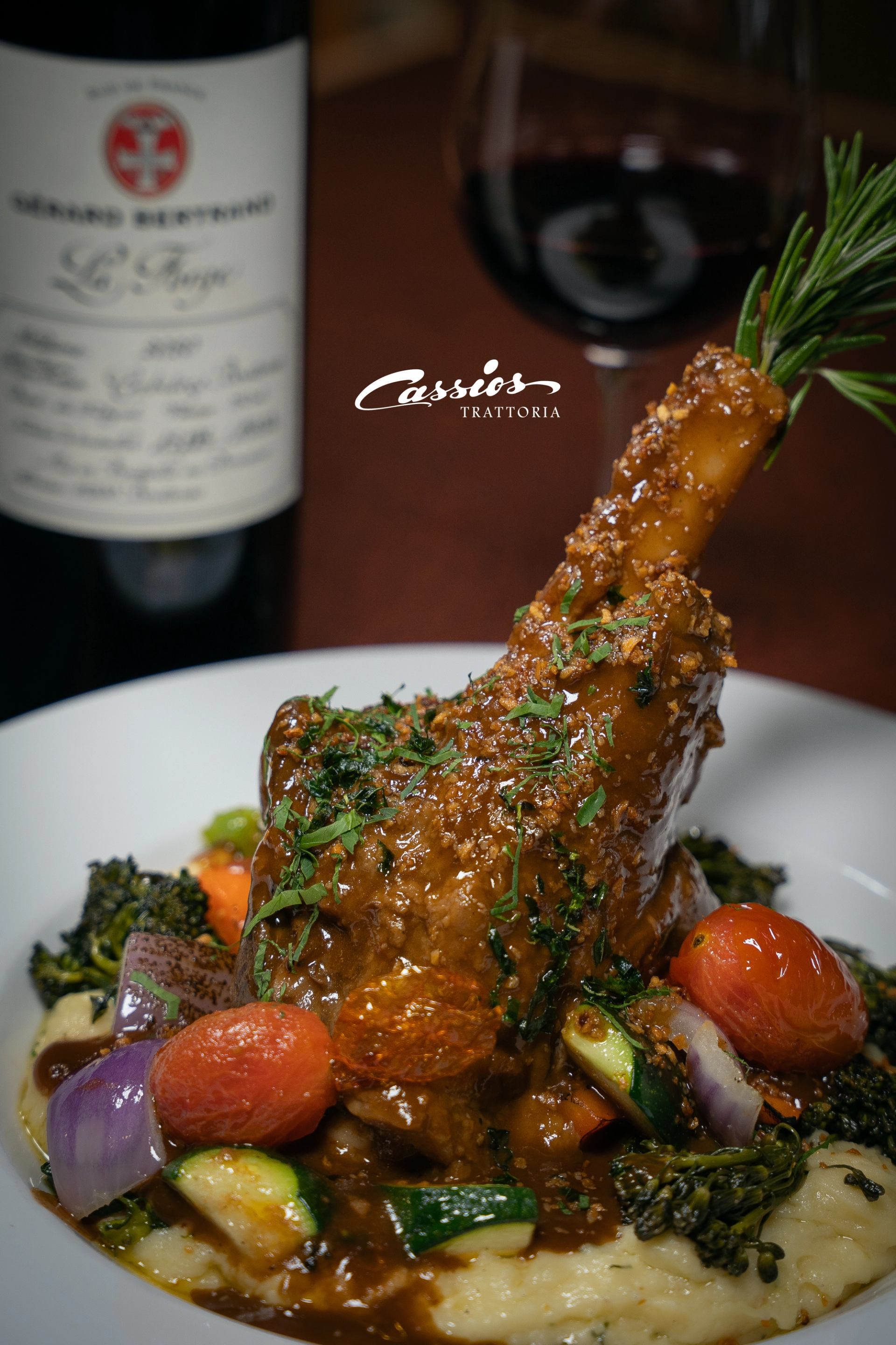 Braised lamb shank on mashed potatoes with vegetables, wine bottle and glass behind.