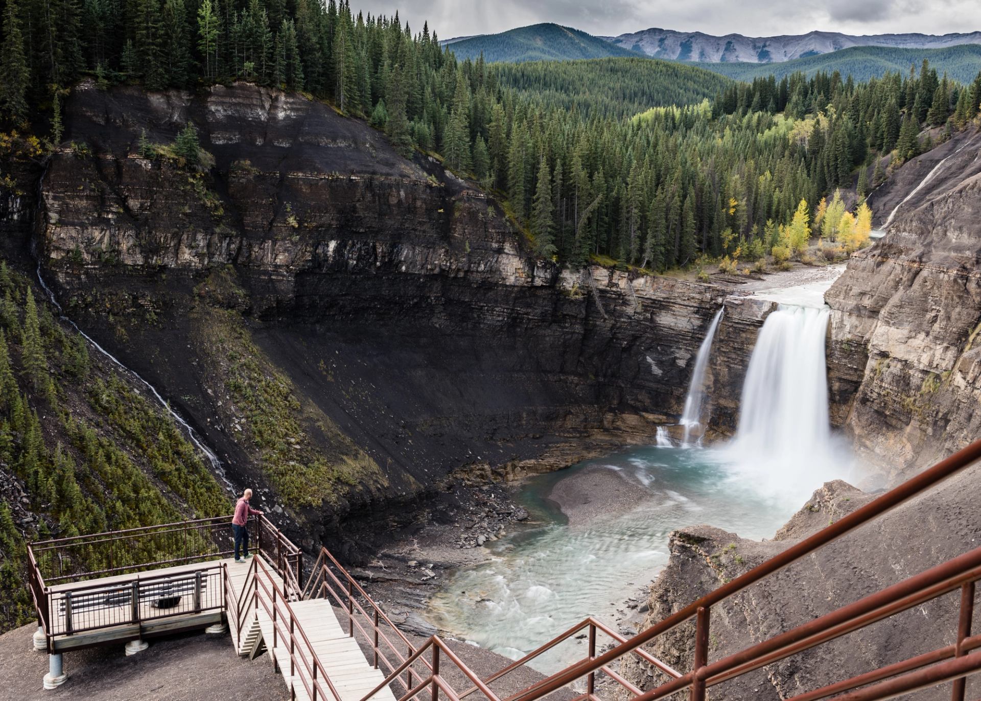 Wooden viewing platform overlooks tall waterfall, rocky canyon walls and dense evergreen forest.