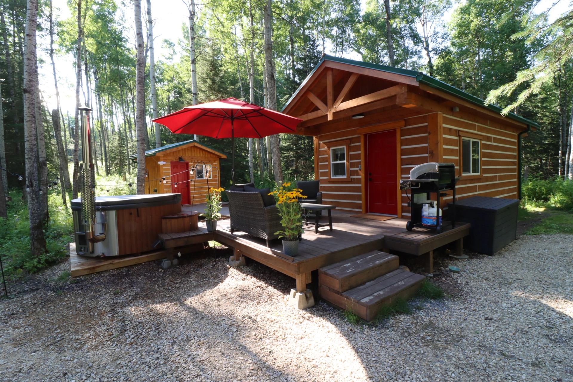 Cozy cabin with deck, hot tub, red umbrella, and grill nestled in a forest.
