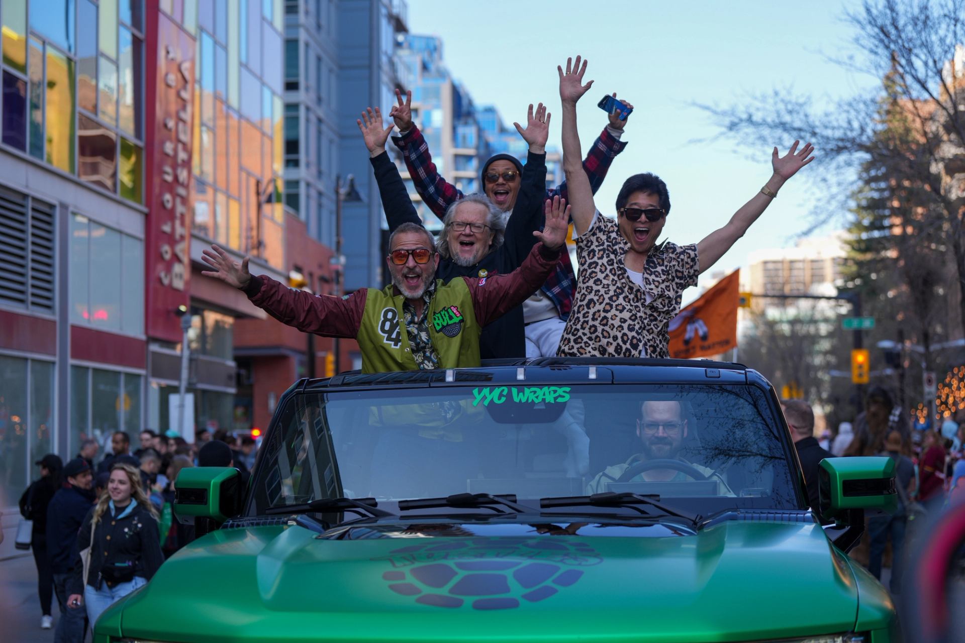 Group of people stand in green convertible on busy city street, raising arms.
