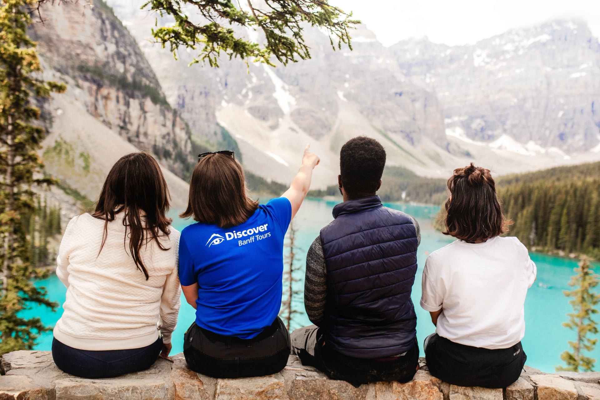 Four people sit on a stone wall overlooking turquoise lake and mountain scenery.