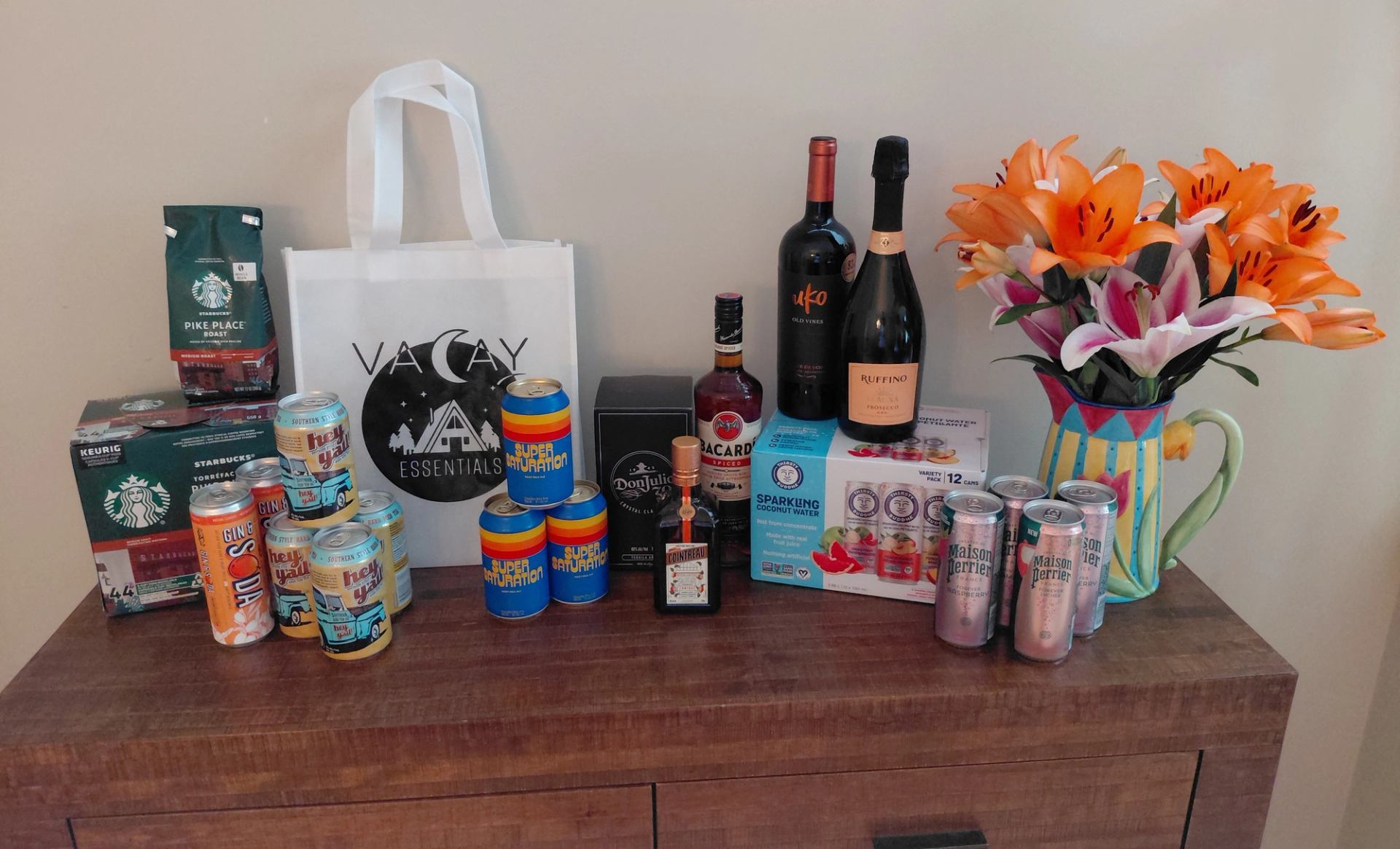 Assorted drinks and flowers with a “VACAY VIBES” tote on a wooden surface.