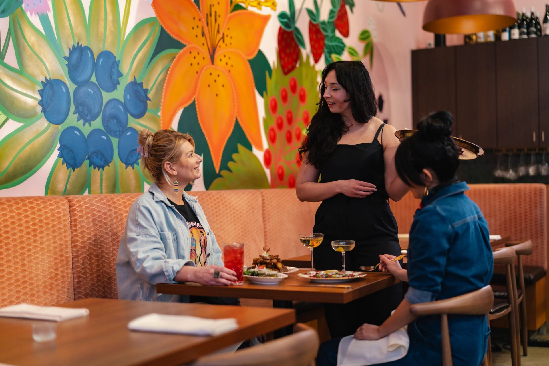 Restaurant server talks with two diners at table beneath colourful fruit and flower mural.
