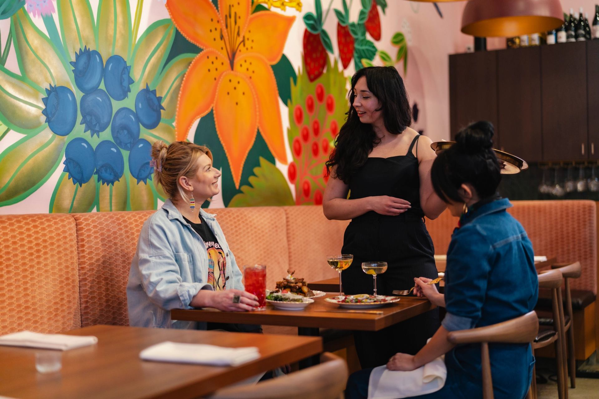 Restaurant server talks with two diners at table beneath colourful fruit and flower mural.