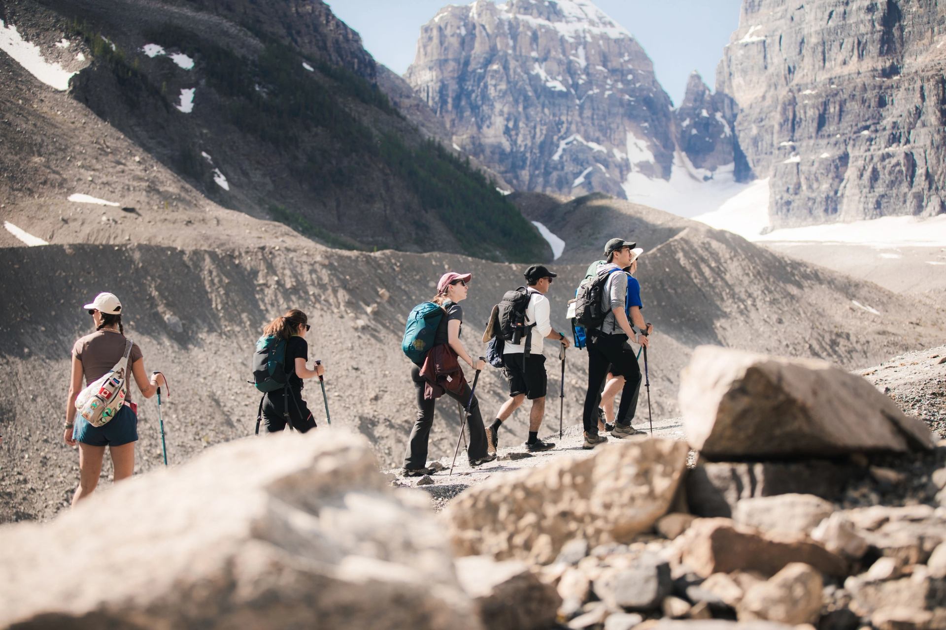 Hikers with backpacks and poles walk along rocky mountain trail below snow-capped peaks.