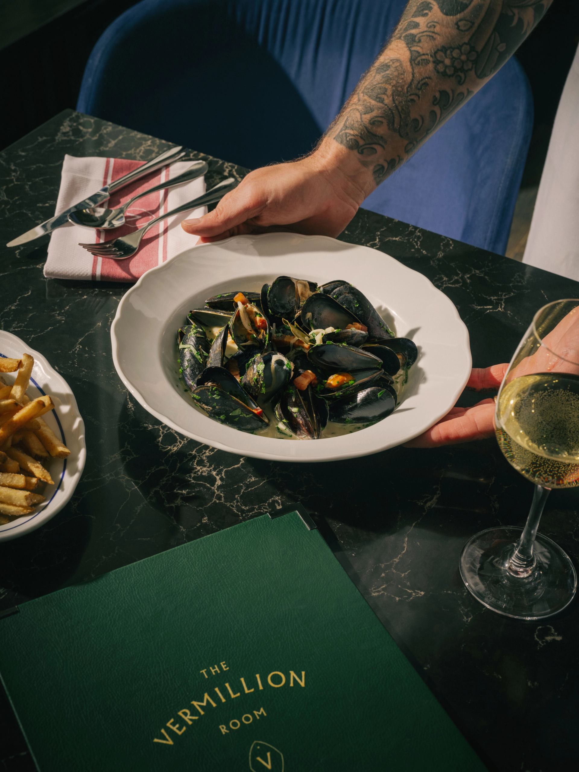 Plate of mussels on a marble table with fries, wine glass, and a green Vermillion Room menu.