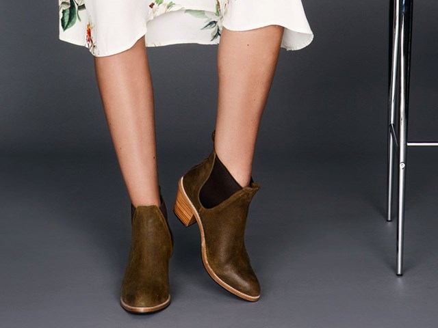 Brown suede ankle boots on display beside a chair, with white dress hems visible.