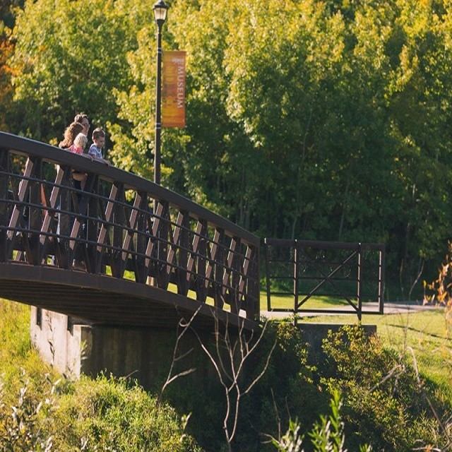 Wooden bridge railing with visitors, Muskoseepi Park sign, and green trees in background.
