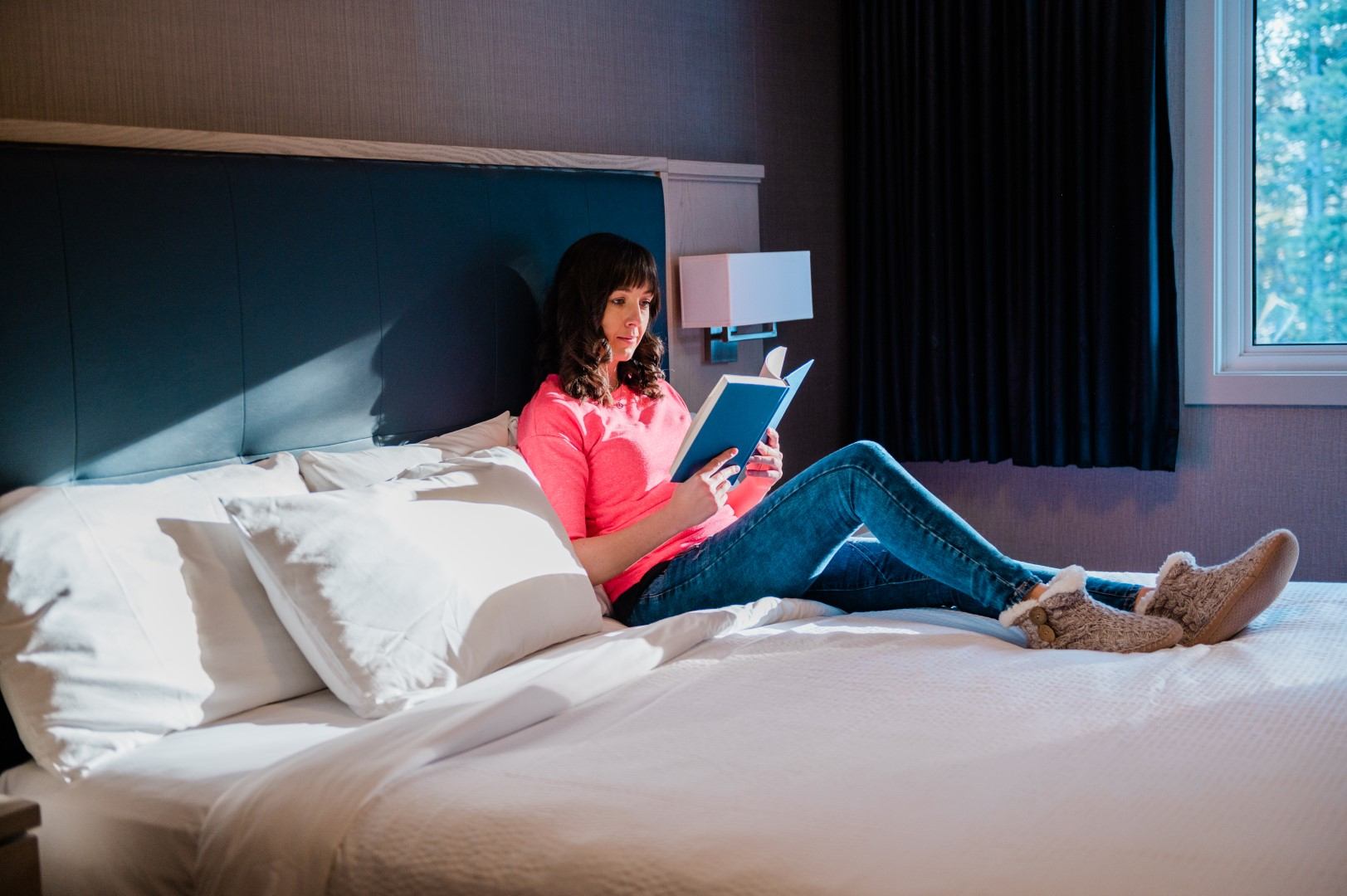 Person relaxing on a bed reading a book in a bright, cozy lodge room.