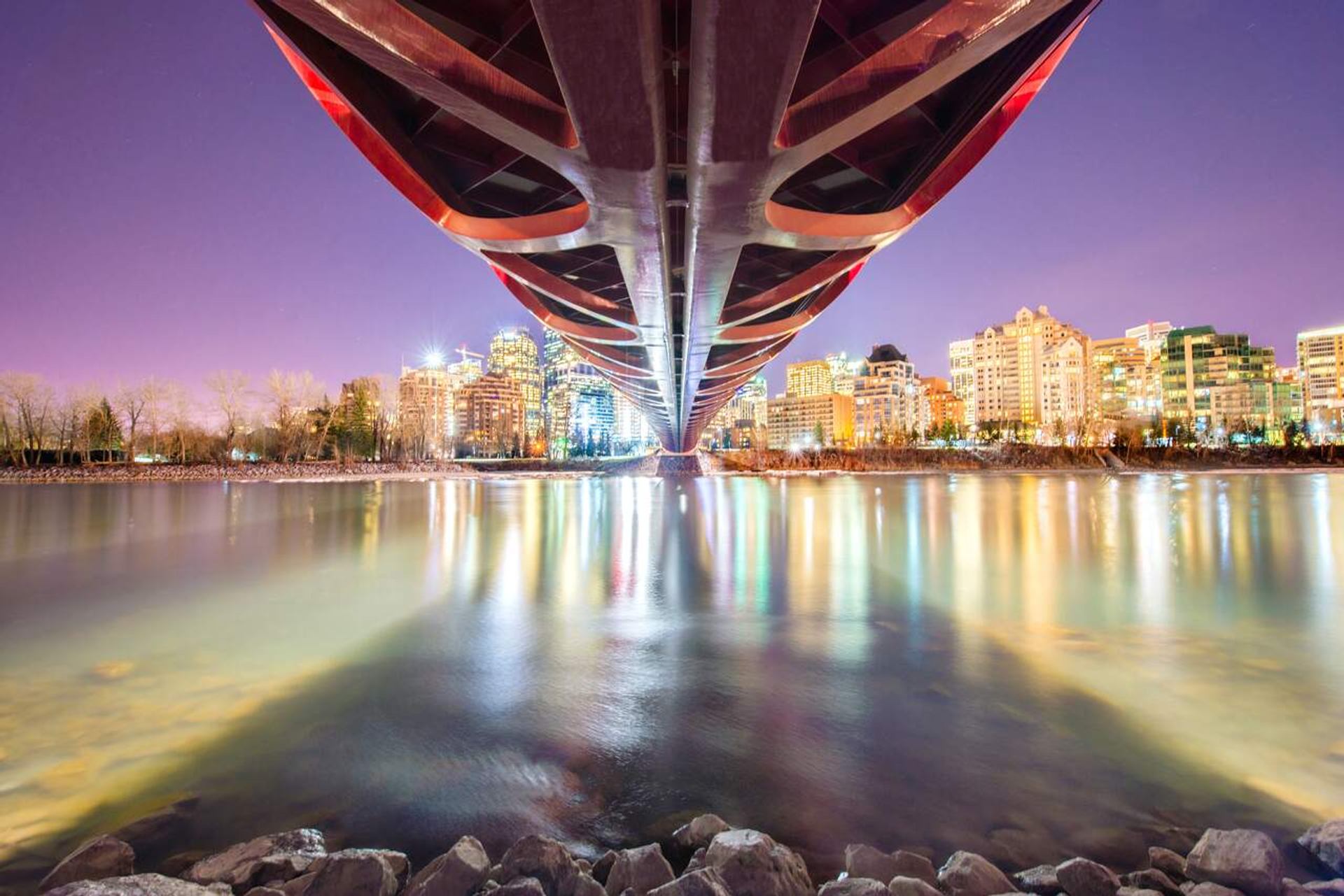 Under-bridge view of illuminated city skyline reflecting on calm river at night.