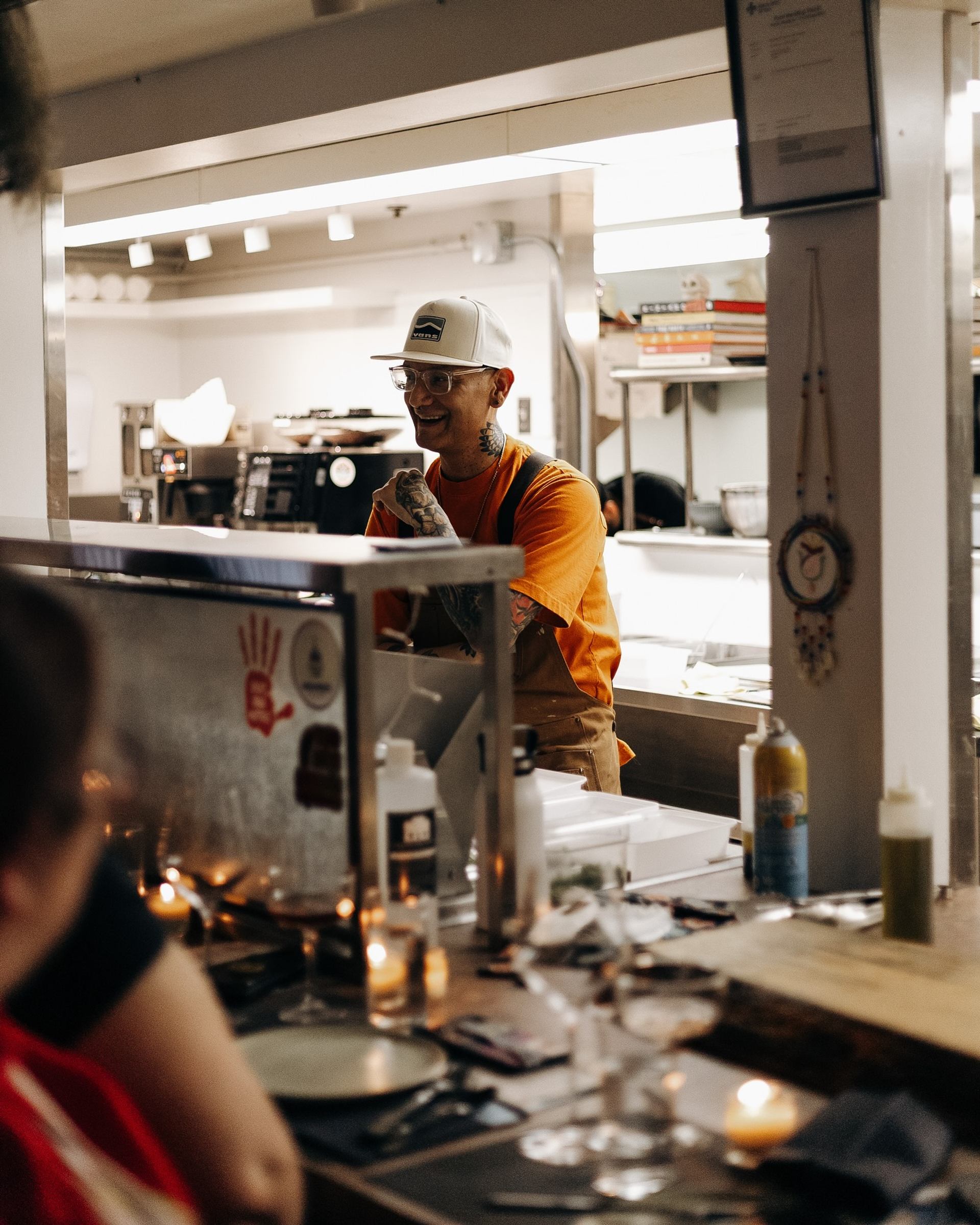 Restaurant kitchen counter with staff member in orange shirt working behind glass partition.