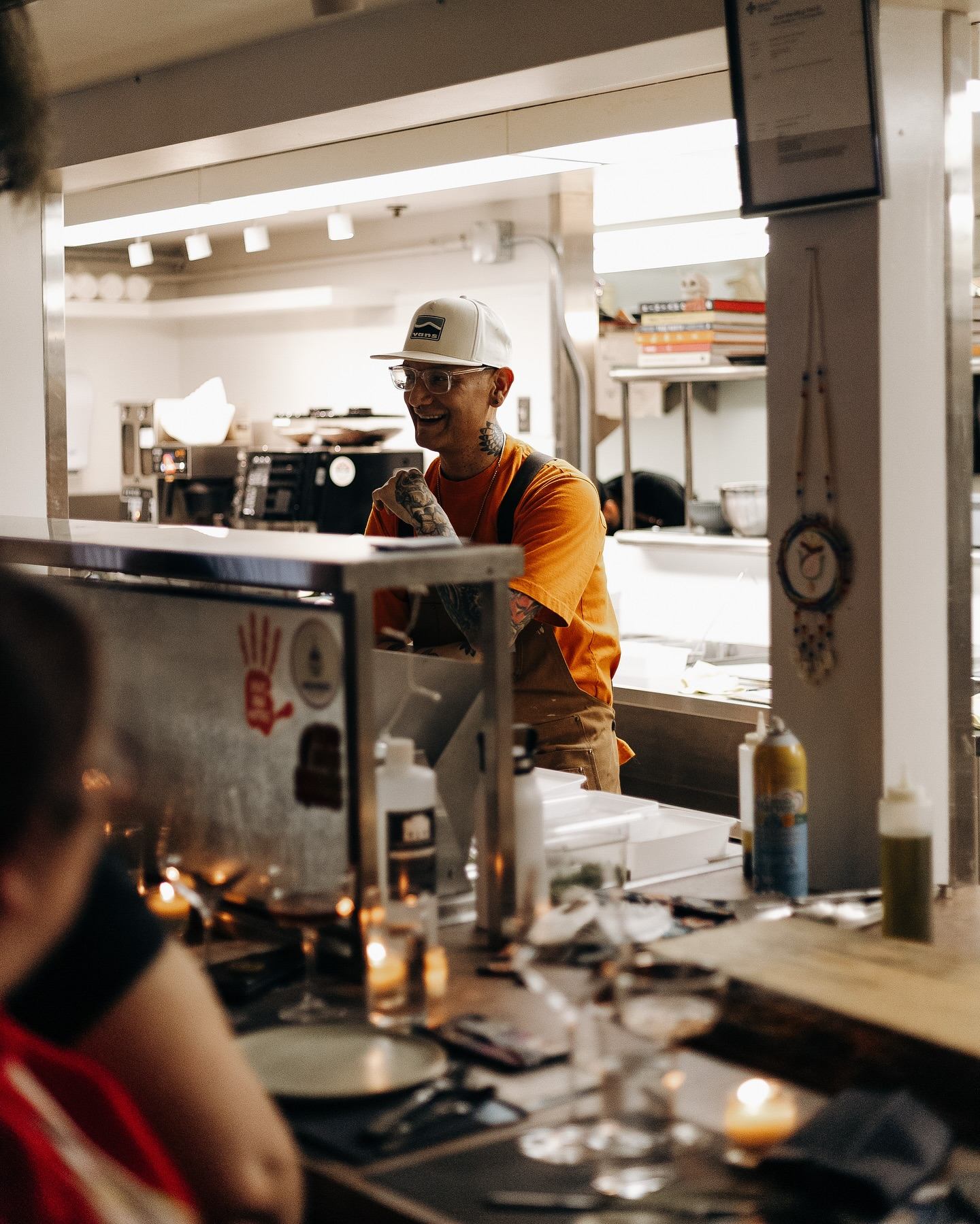 Restaurant kitchen counter with staff member in orange shirt working behind glass partition.