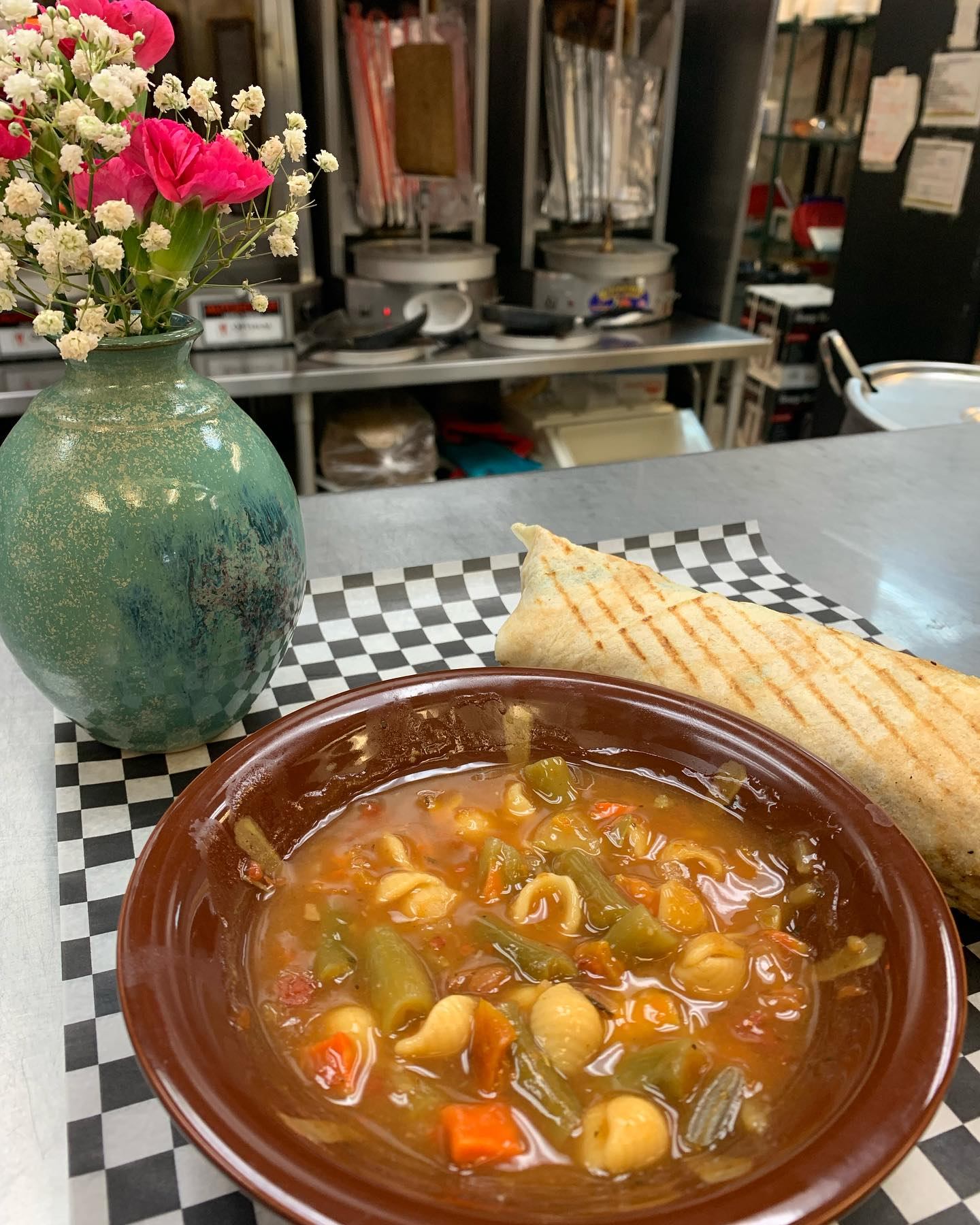 Soup bowl and grilled flatbread on a checkered table, with a floral vase nearby.