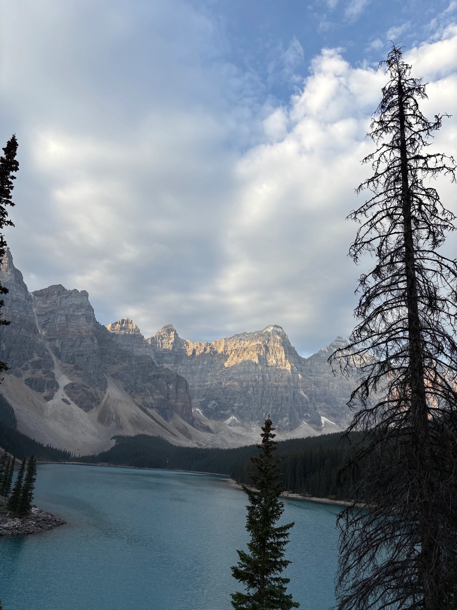 Turquoise mountain lake with evergreen trees, rocky peaks and partly cloudy sky.