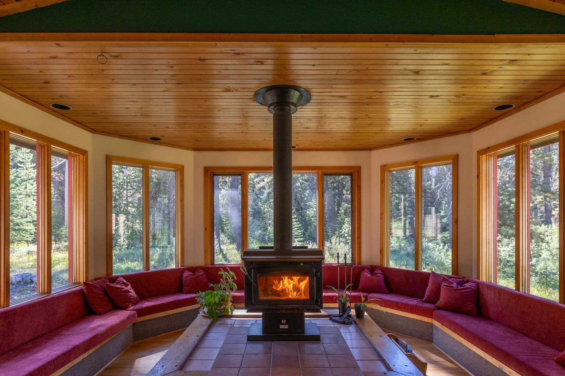 Common room with wraparound windows overlooking forest, central wood-burning stove.