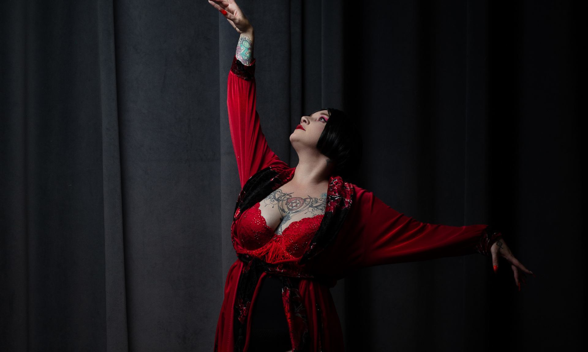 Performer in red robe and lingerie poses with outstretched arms against dark curtain backdrop.