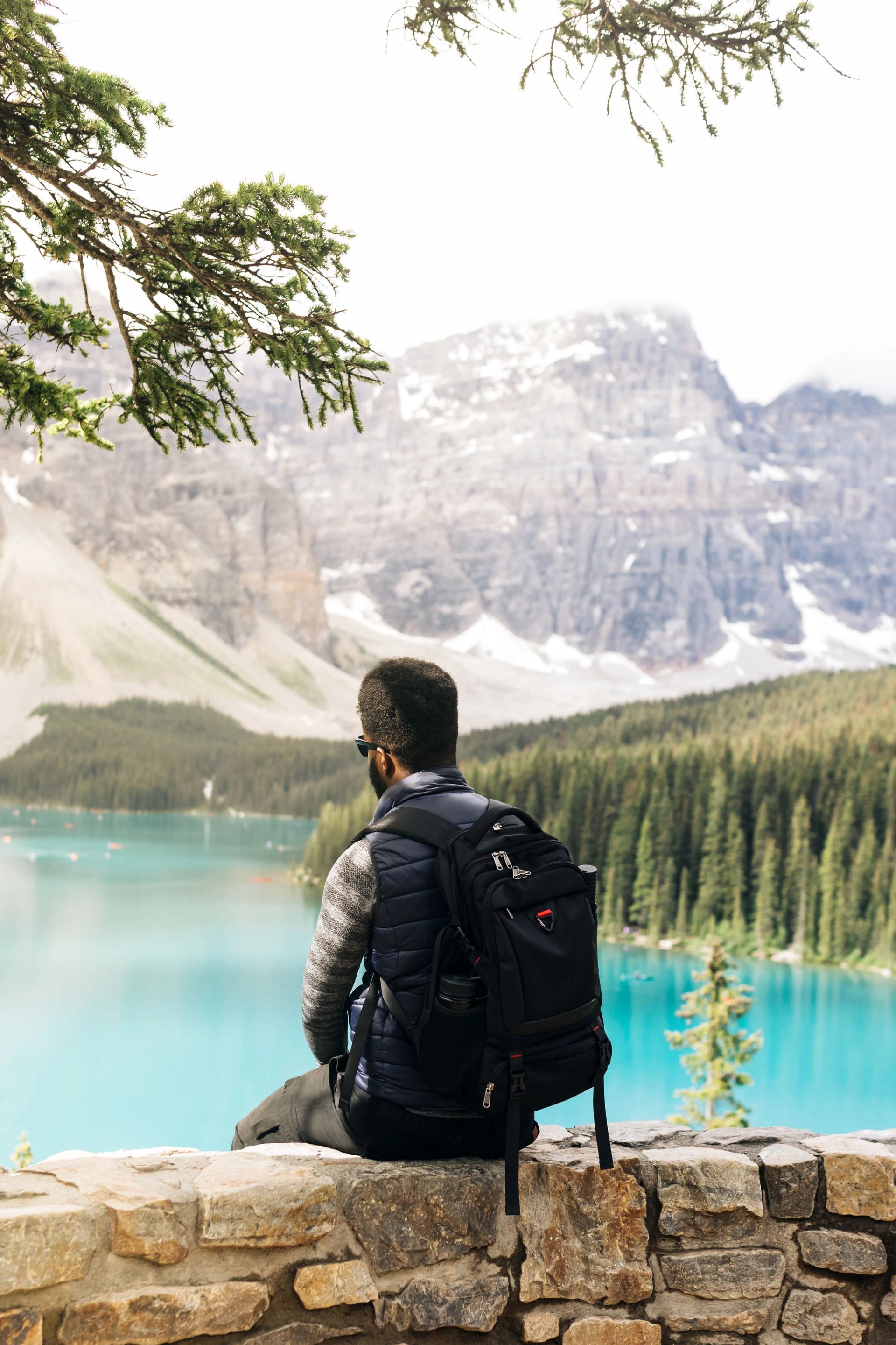 Person with backpack sitting on stone wall overlooking turquoise lake and mountains.