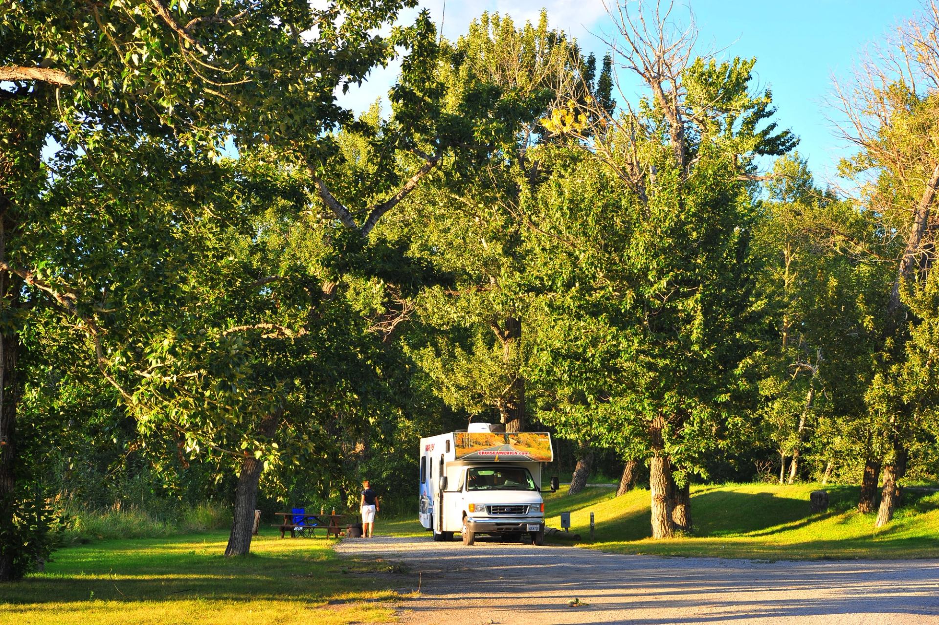 RV parked in shaded campground with trees and two people nearby.