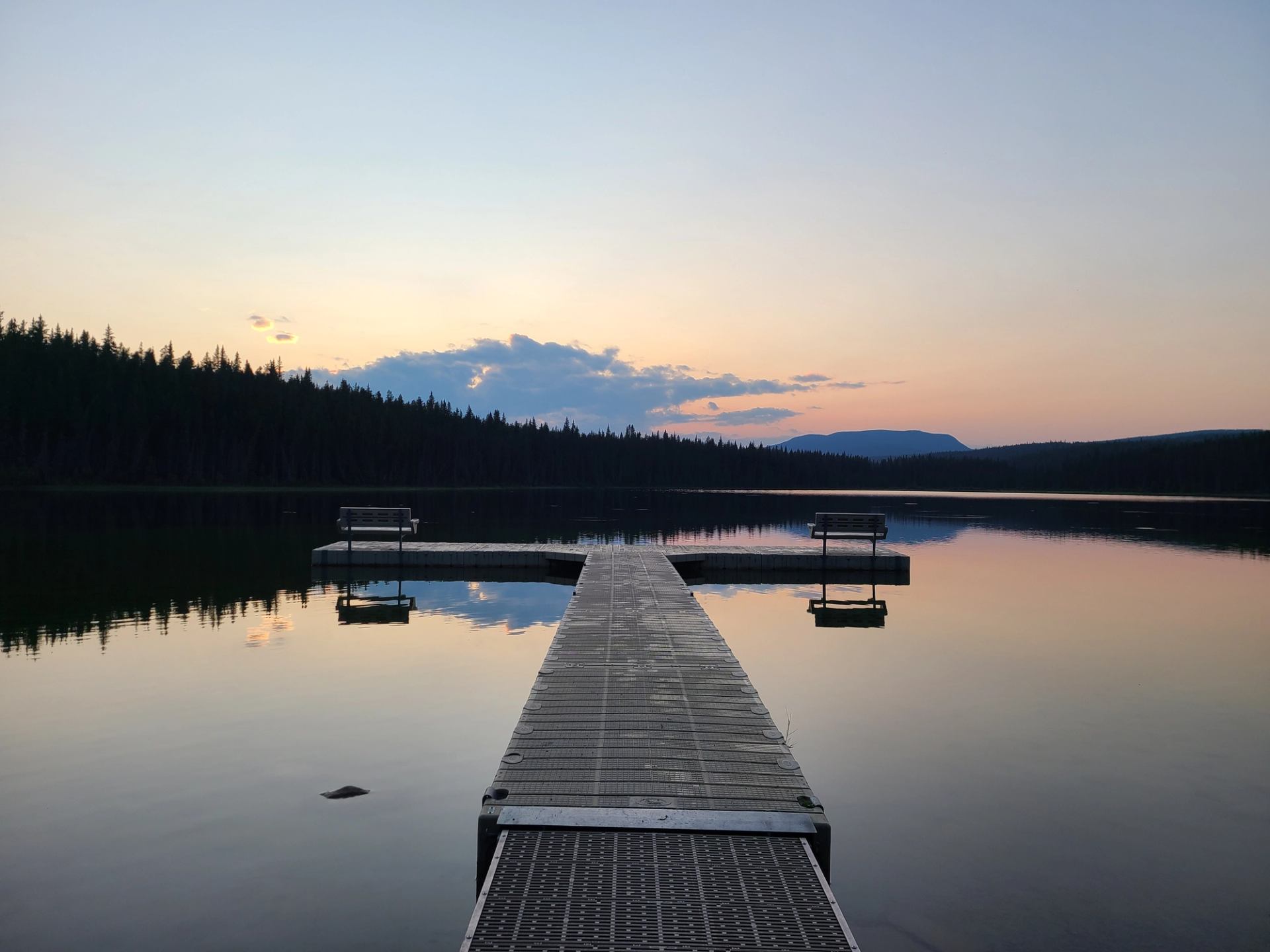 Wooden dock with benches over calm lake, forested shoreline and distant hills.