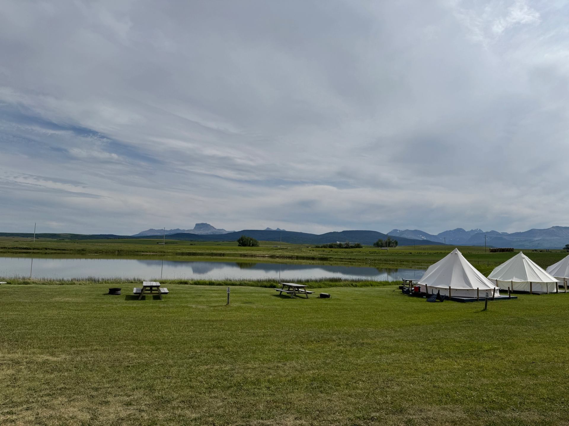 White glamping tents set on grassy fields beside a still lake and mountain backdrop.