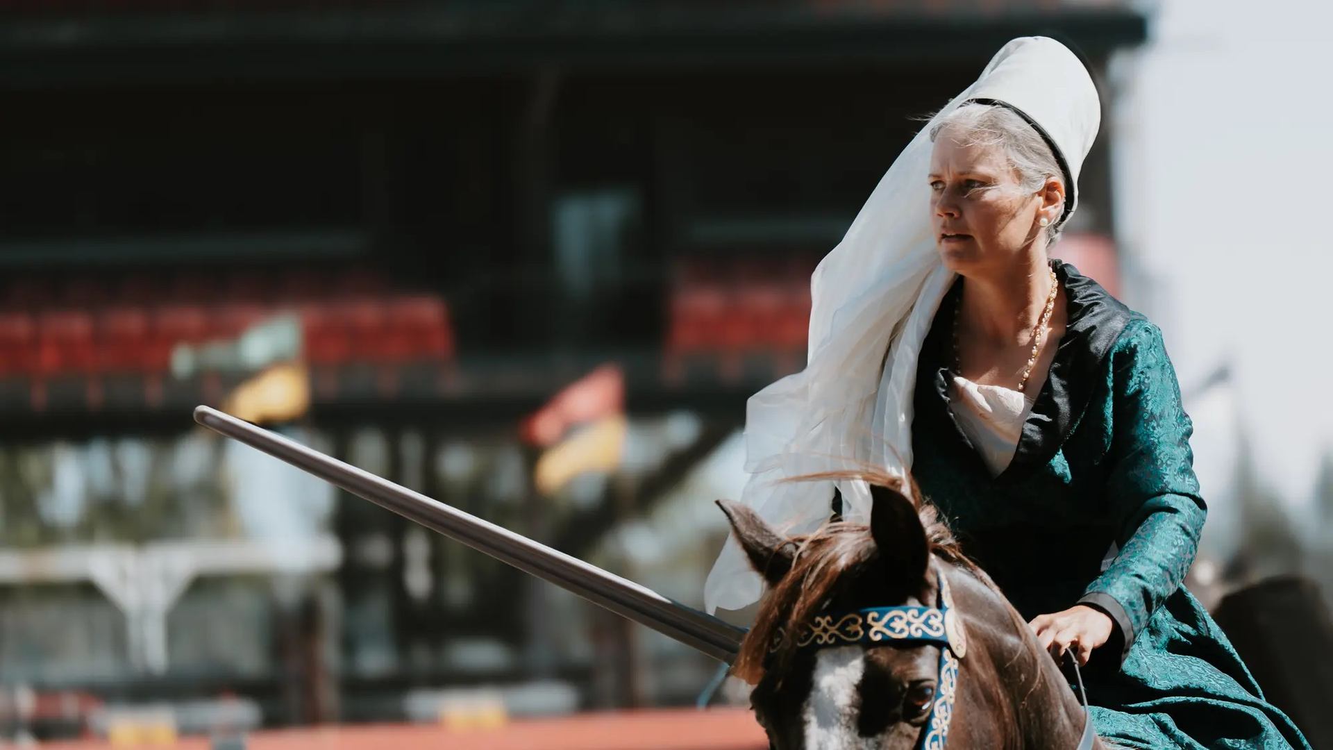 Rider in green medieval dress on horseback with lance at outdoor jousting arena.