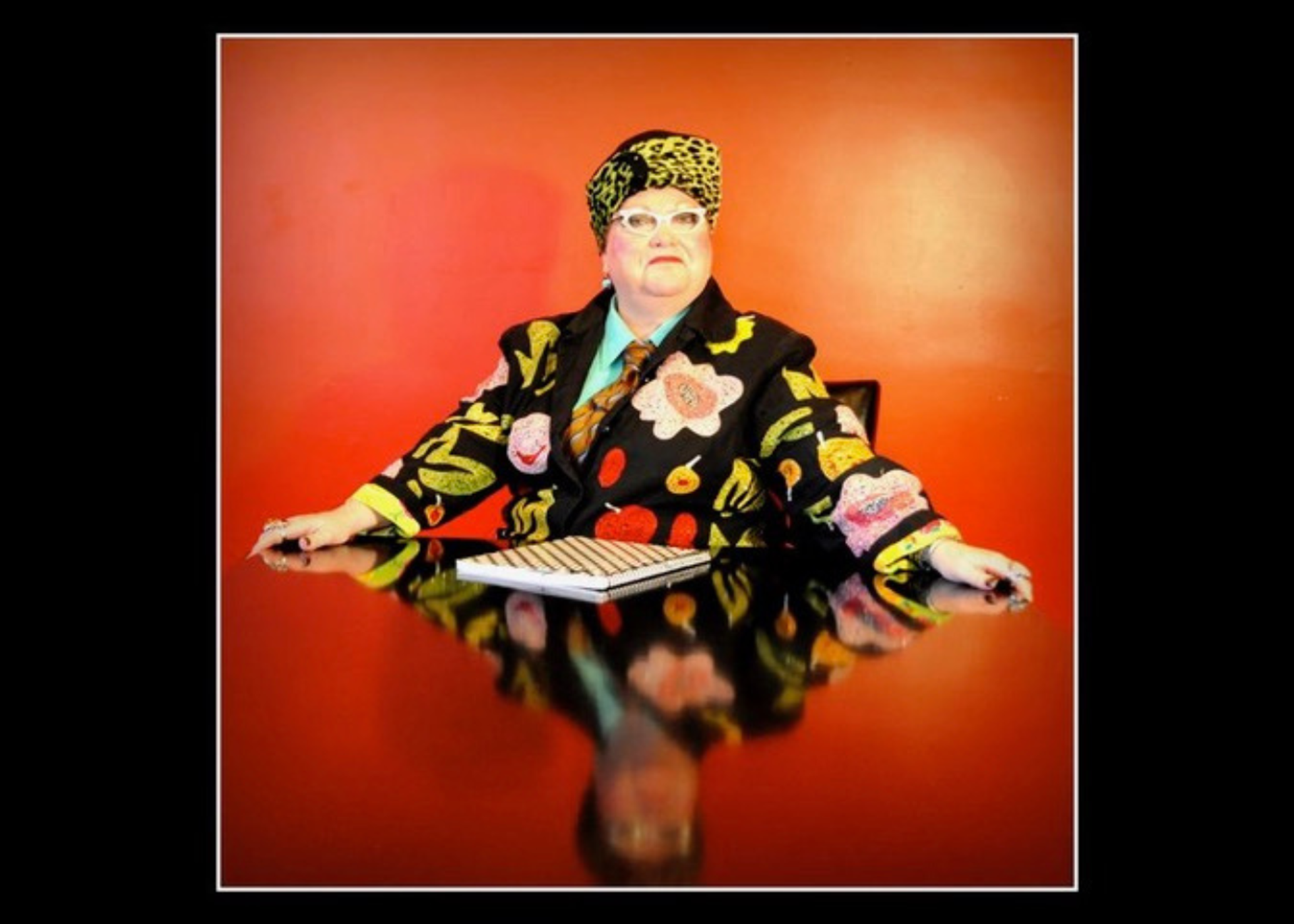 Person in patterned suit and headscarf sits at glossy table against red wall.
