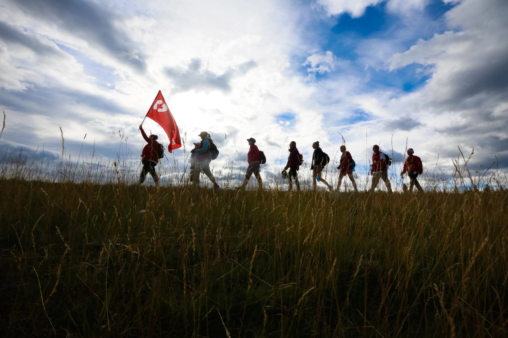 A silhouetted group of hikers walks along a grassy ridge, the leader holding a red flag, under a dramatic cloudy sky.