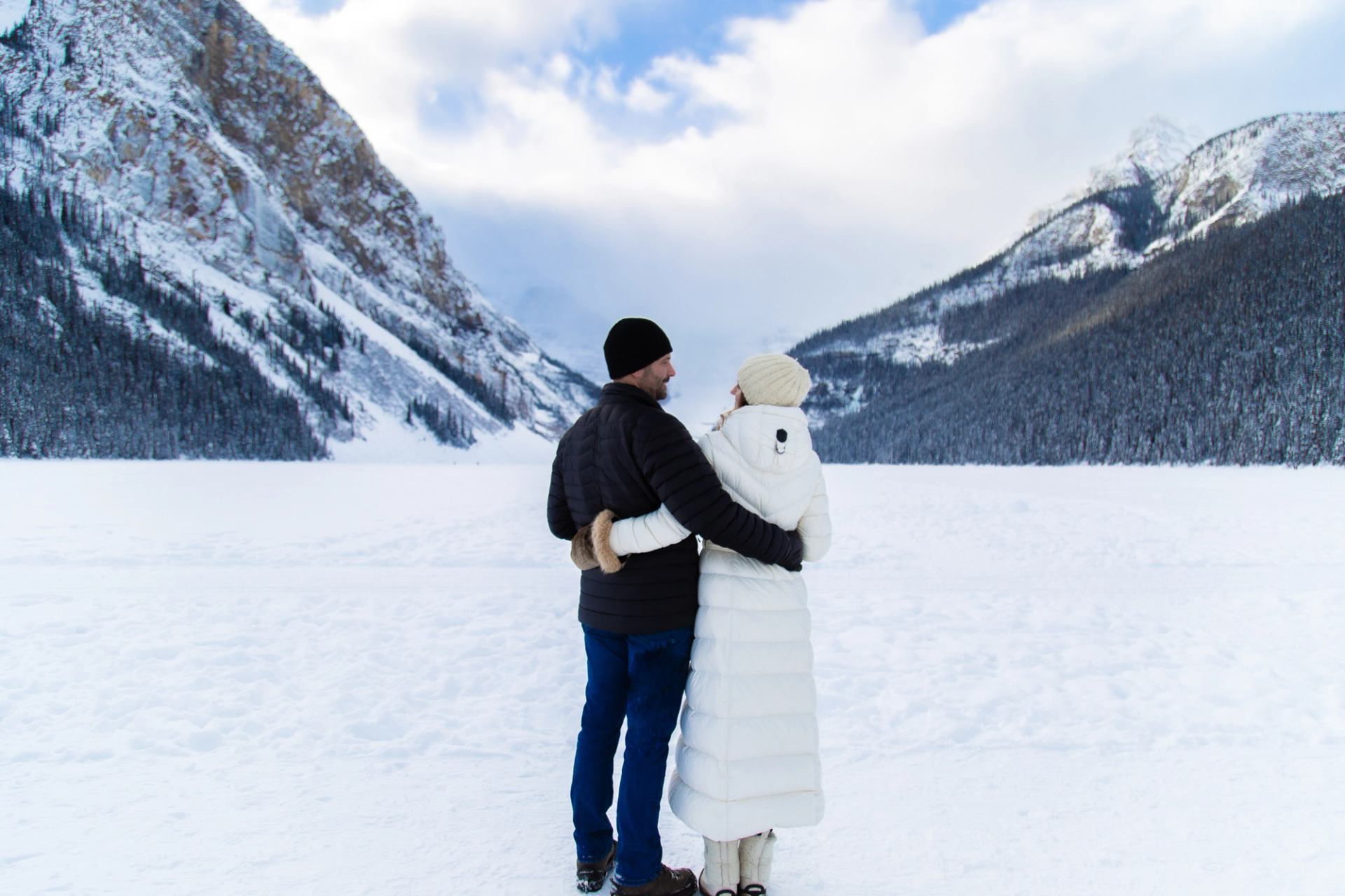 Two people standing arm in arm on a frozen lake surrounded by snow-covered mountains