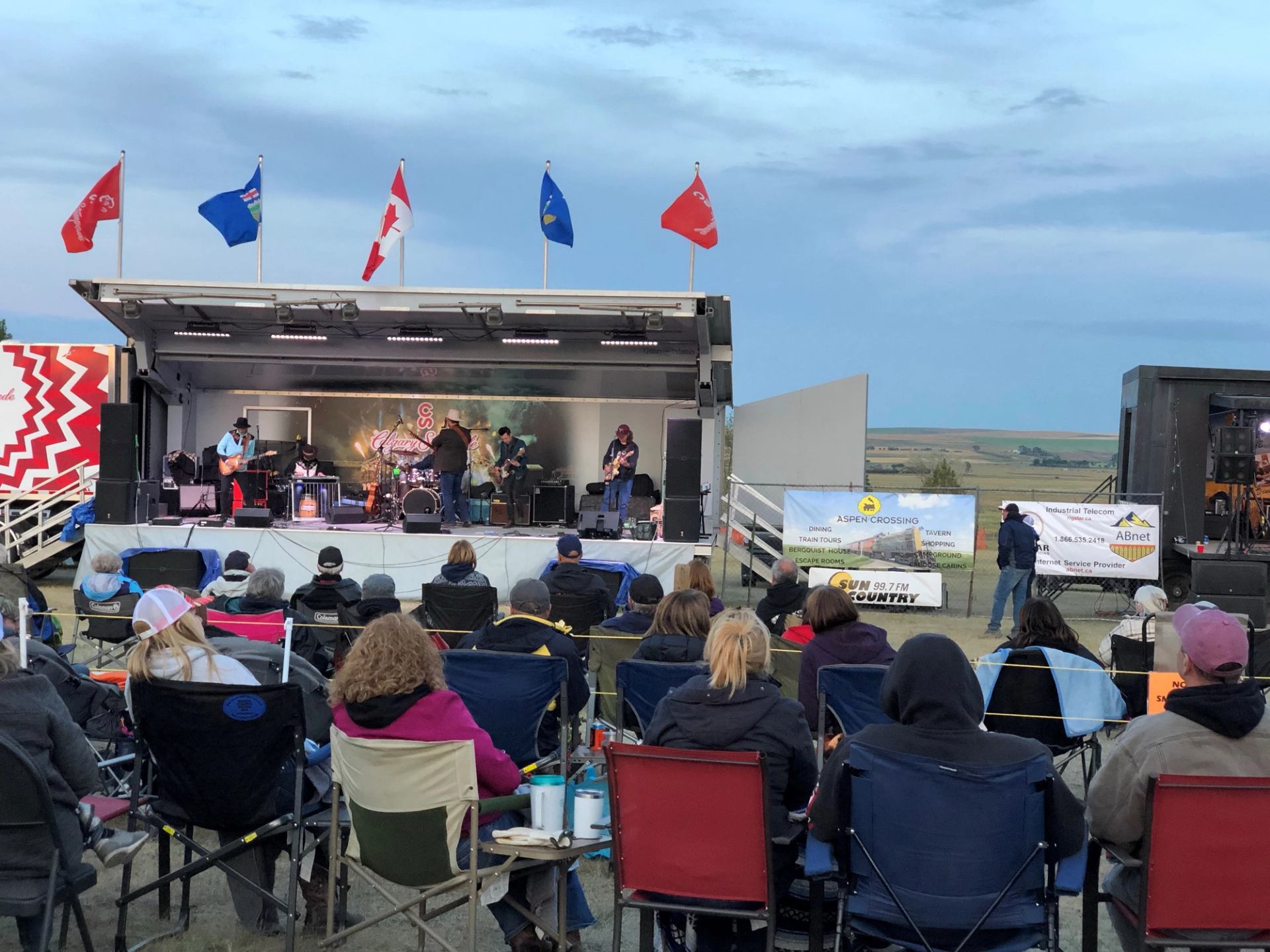 Outdoor audience in camp chairs watching live band perform on trailer stage with flags.
