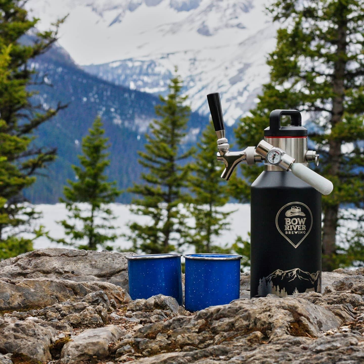 Bow River Brewing growler with tap beside blue mugs on rocks, snowy mountains behind.