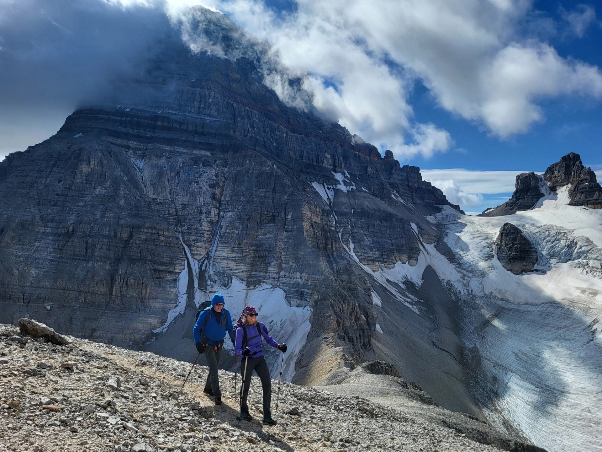 Two hikers on rocky trail with towering mountain and patchy clouds in background.