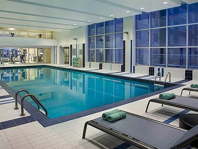 Indoor pool with lounge chairs, handrails, and gym visible through windows.