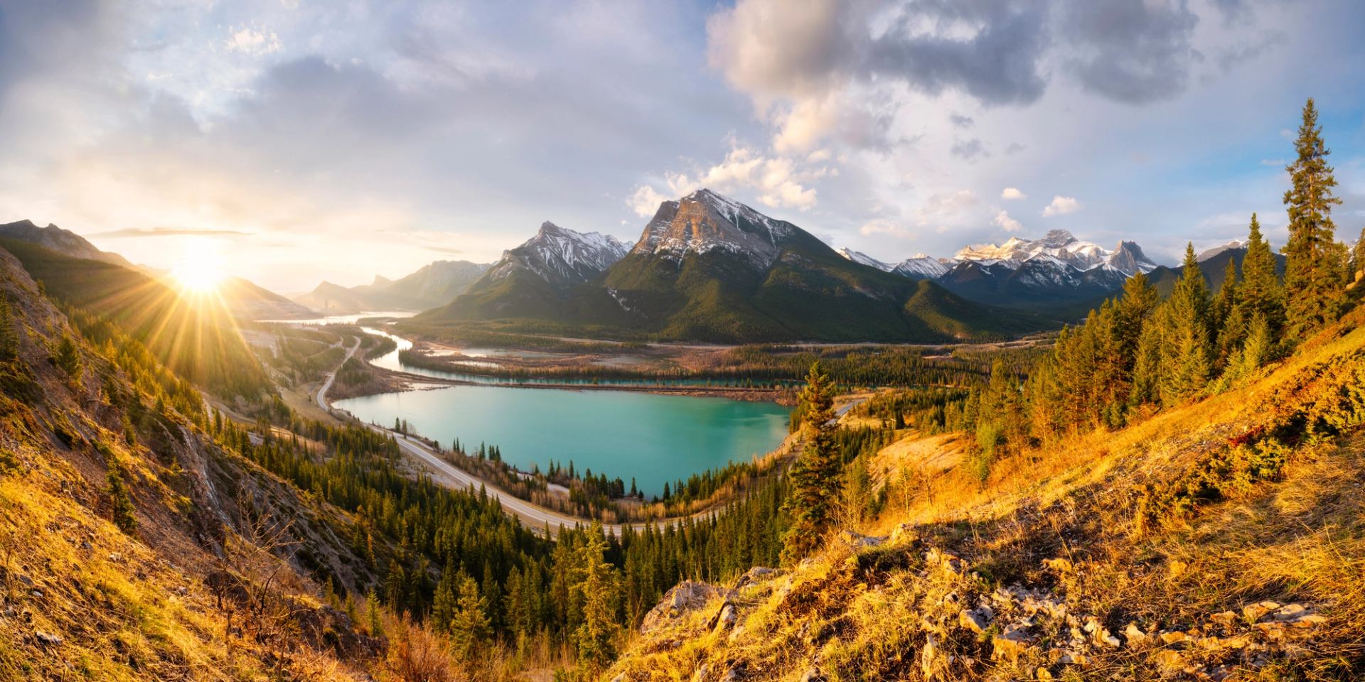 Mountain lake with turquoise water, winding highway and sunlit forested slopes under clouds.