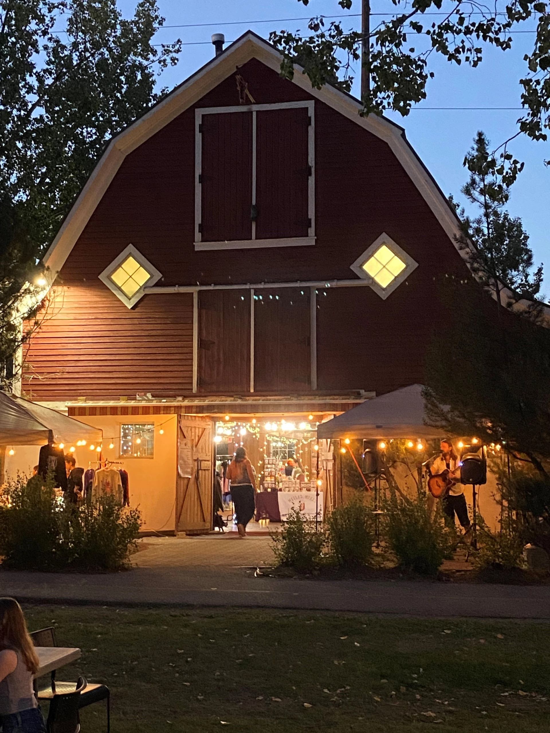 Red barn glowing at dusk with string lights and artisan vendors set up outside.