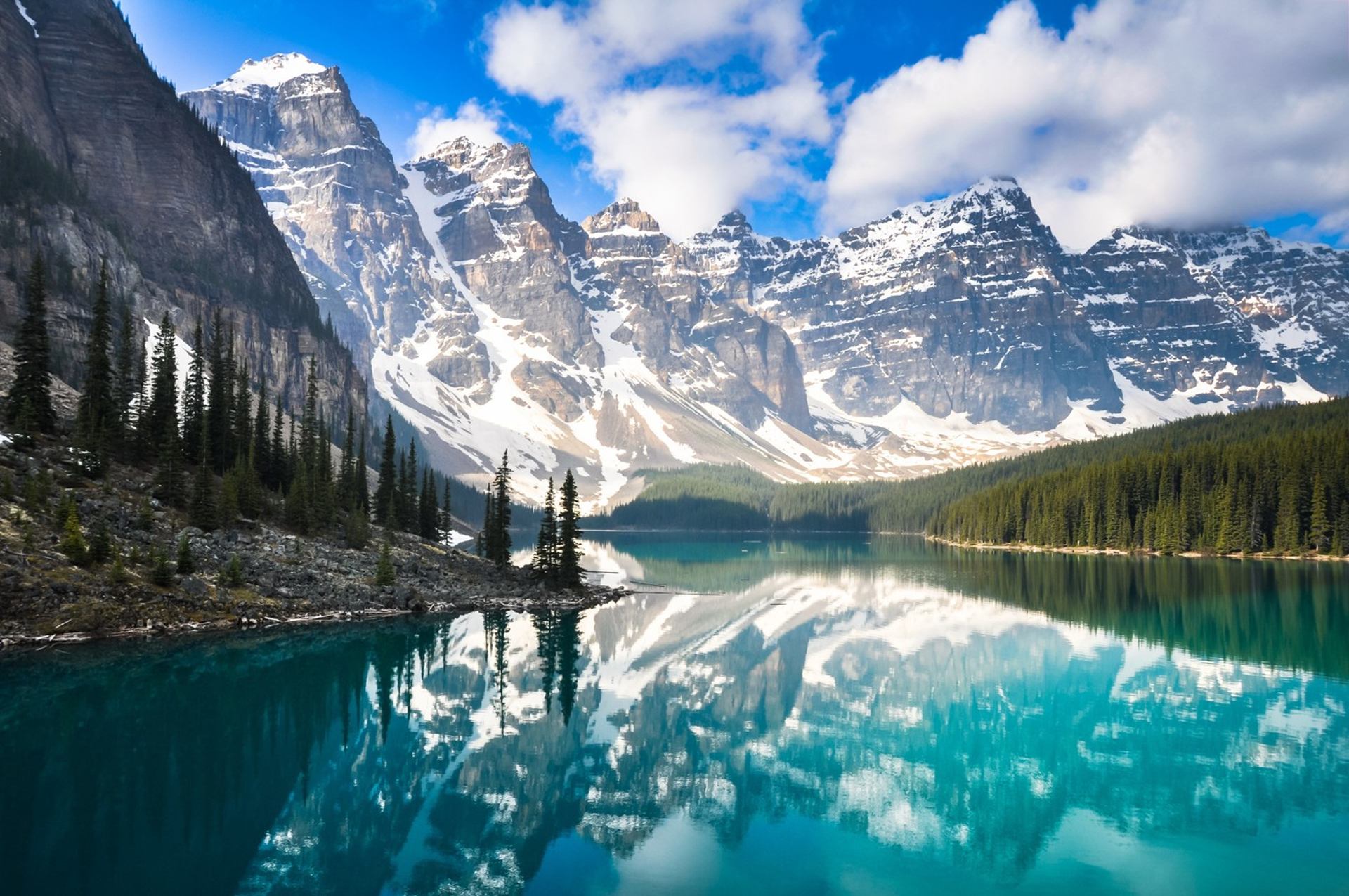 Turquoise lake reflecting snow-capped mountains and pine trees under a blue sky.