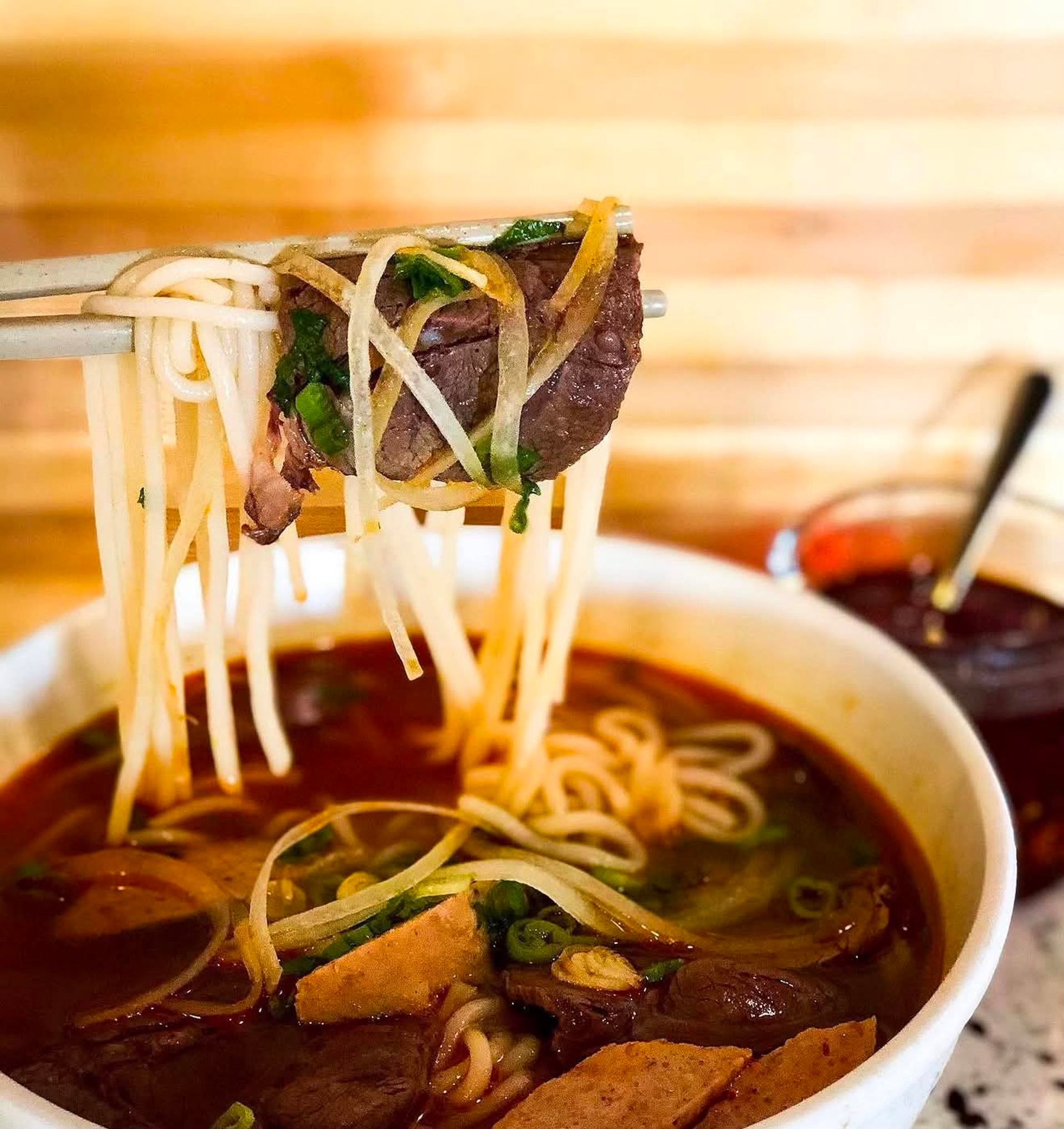Chopsticks lift rice noodles and beef from a bowl of red broth noodle soup.