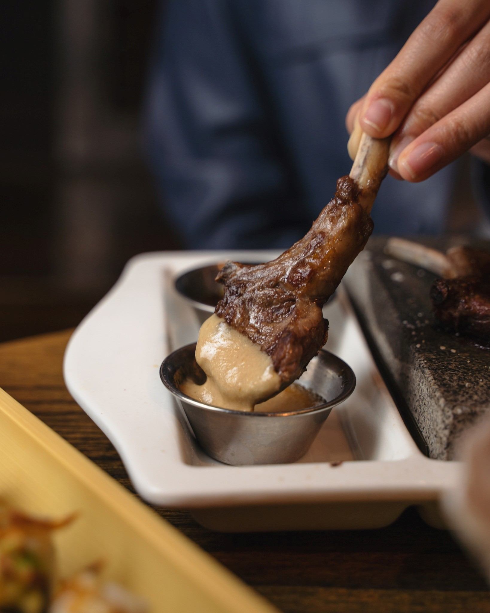 Hand holds grilled sausage over metal dipping cup on white platter.
