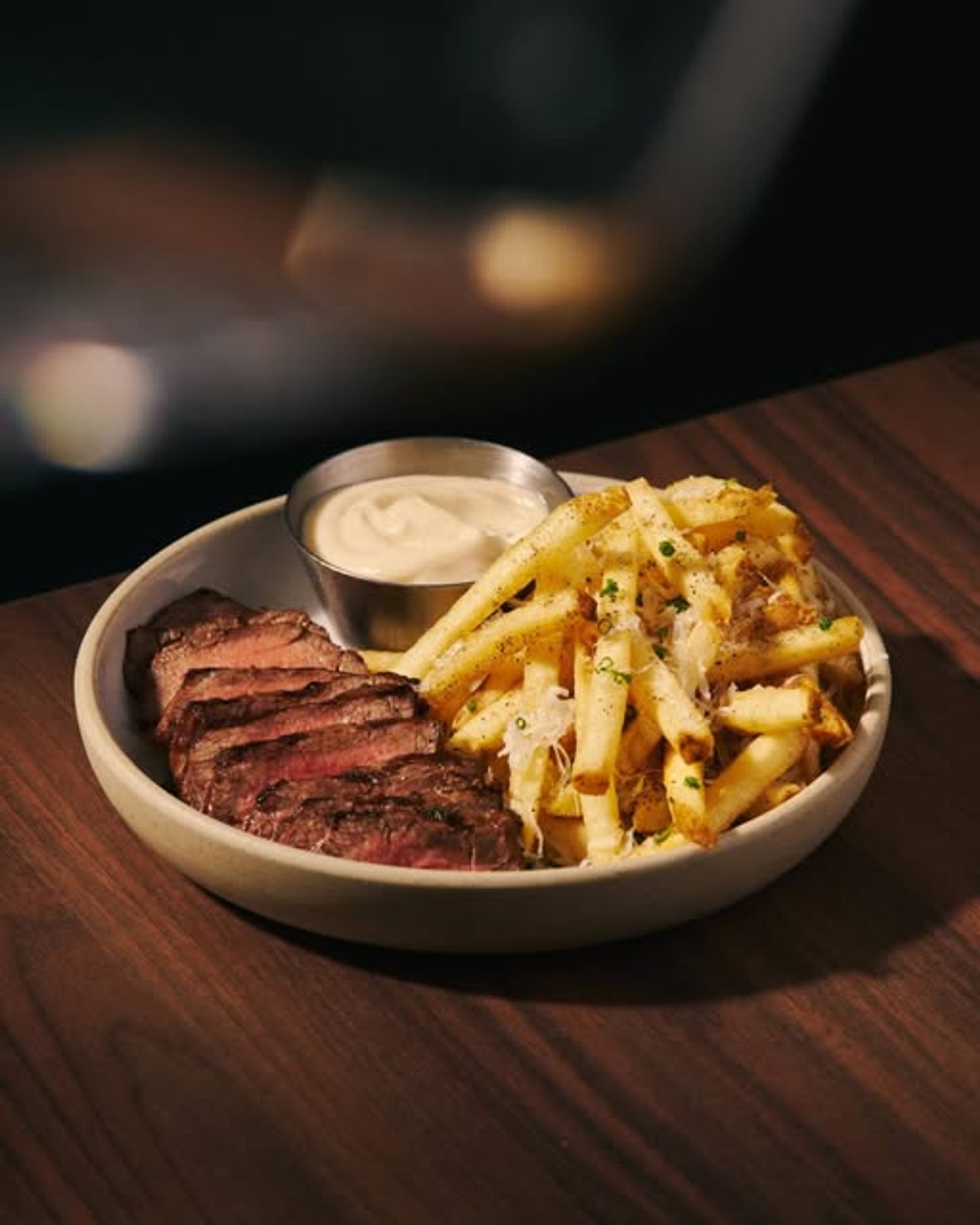 Sliced steak is served beside golden fries, creamy dipping sauce, and melted cheese on a wooden table.