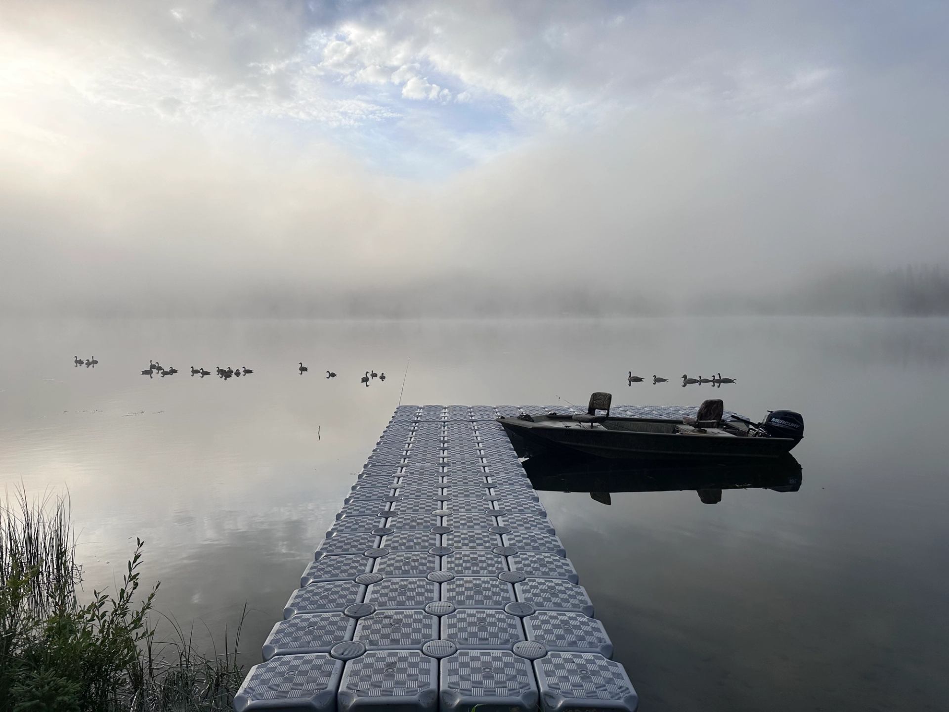 Floating dock with small motorboat on calm foggy lake, ducks swimming in distance.