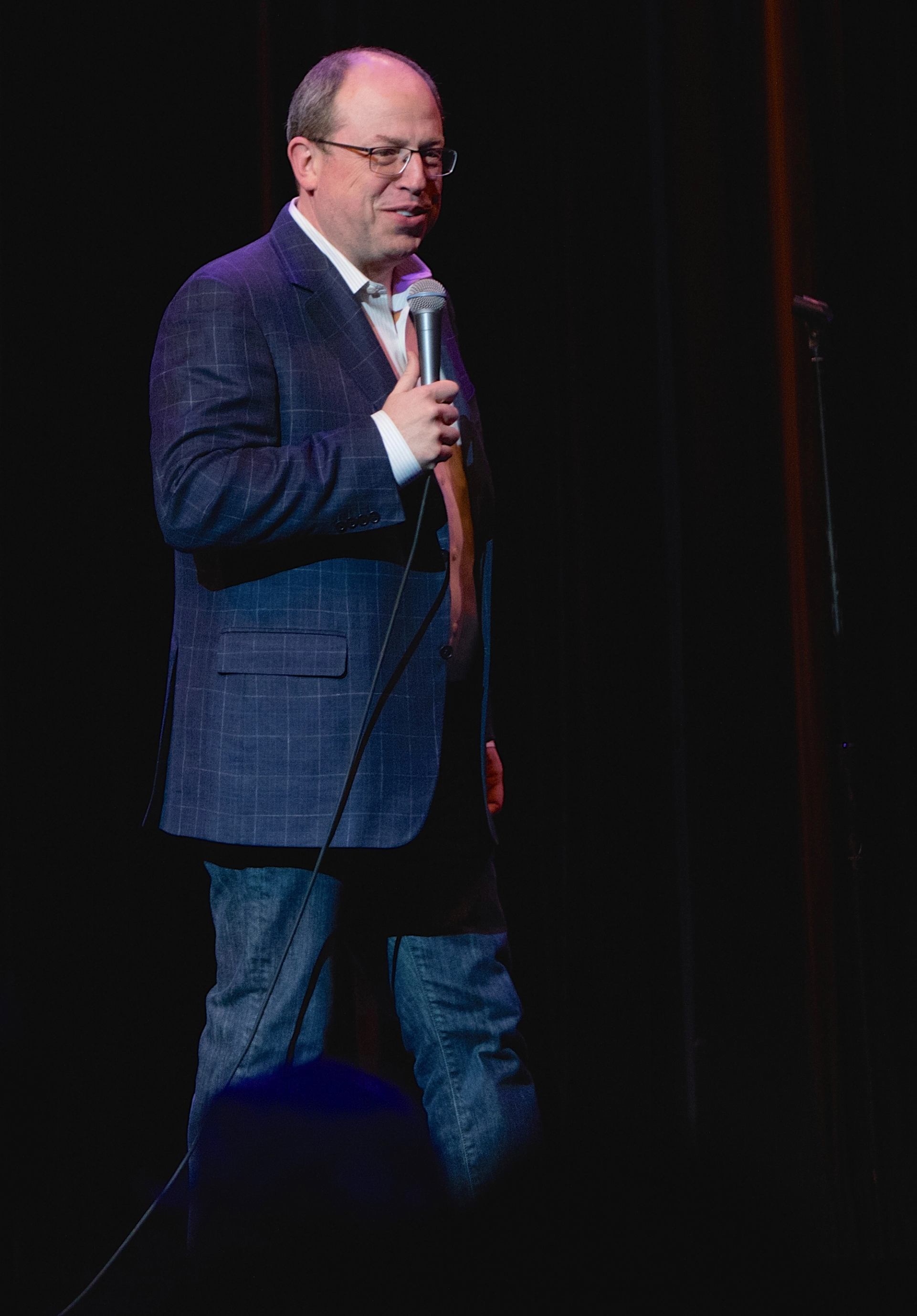 Comedian in blazer and jeans holds a microphone on a dark theatre stage.