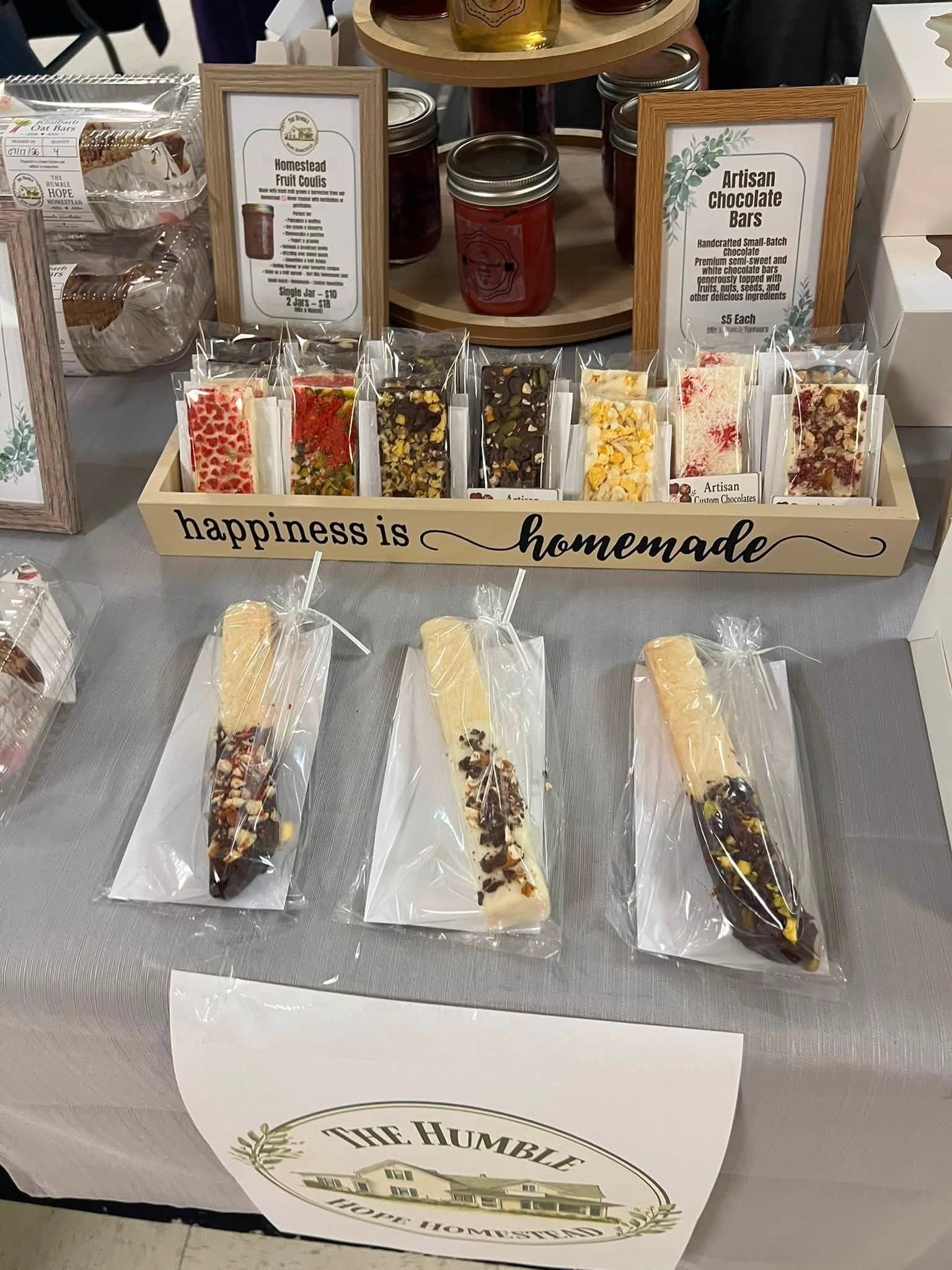 Market table with packaged artisan chocolate bars, dipped treats, jars and framed signs.