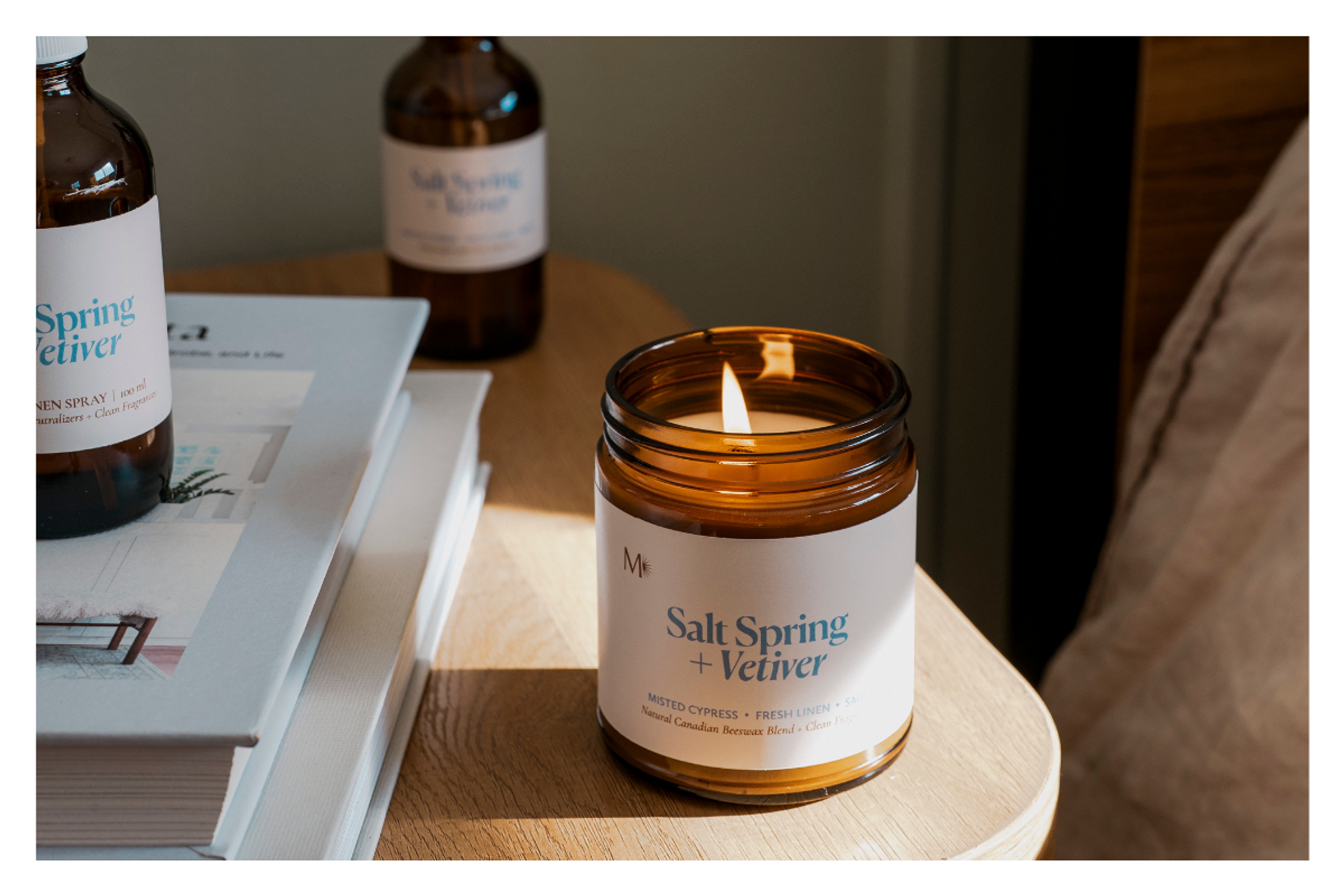 Lit Salt Spring + Vetiver candle on bedside table beside books and linen spray bottles.