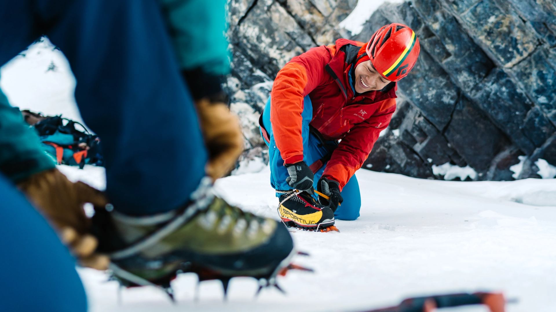 Two climbers adjusting crampons on their boots in the snow, one smiling.