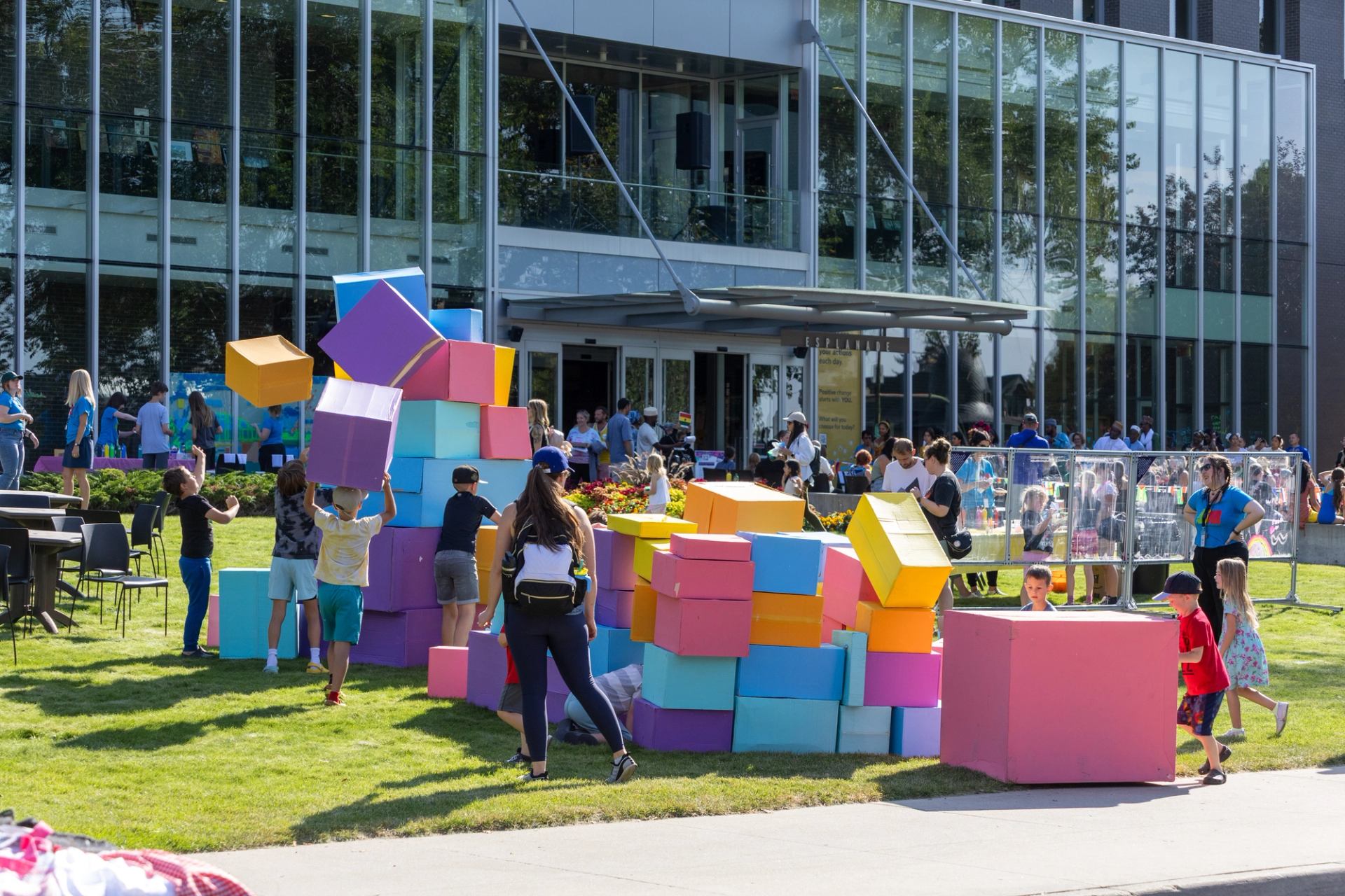 Children and adults stack large colourful foam cubes on grass outside glass building.