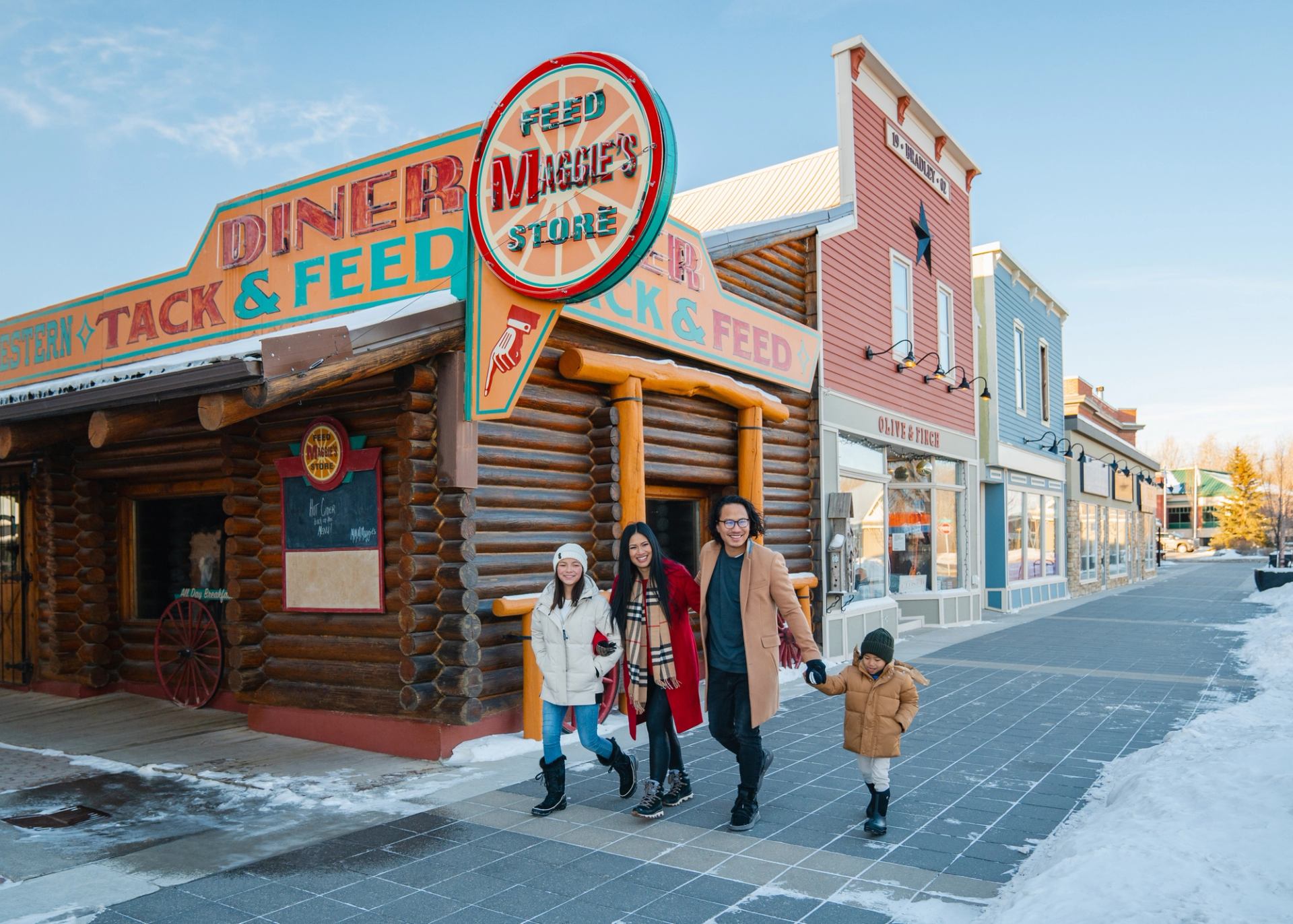 People walk past Maggie’s Feed Store and colourful wooden storefronts on a snowy street.