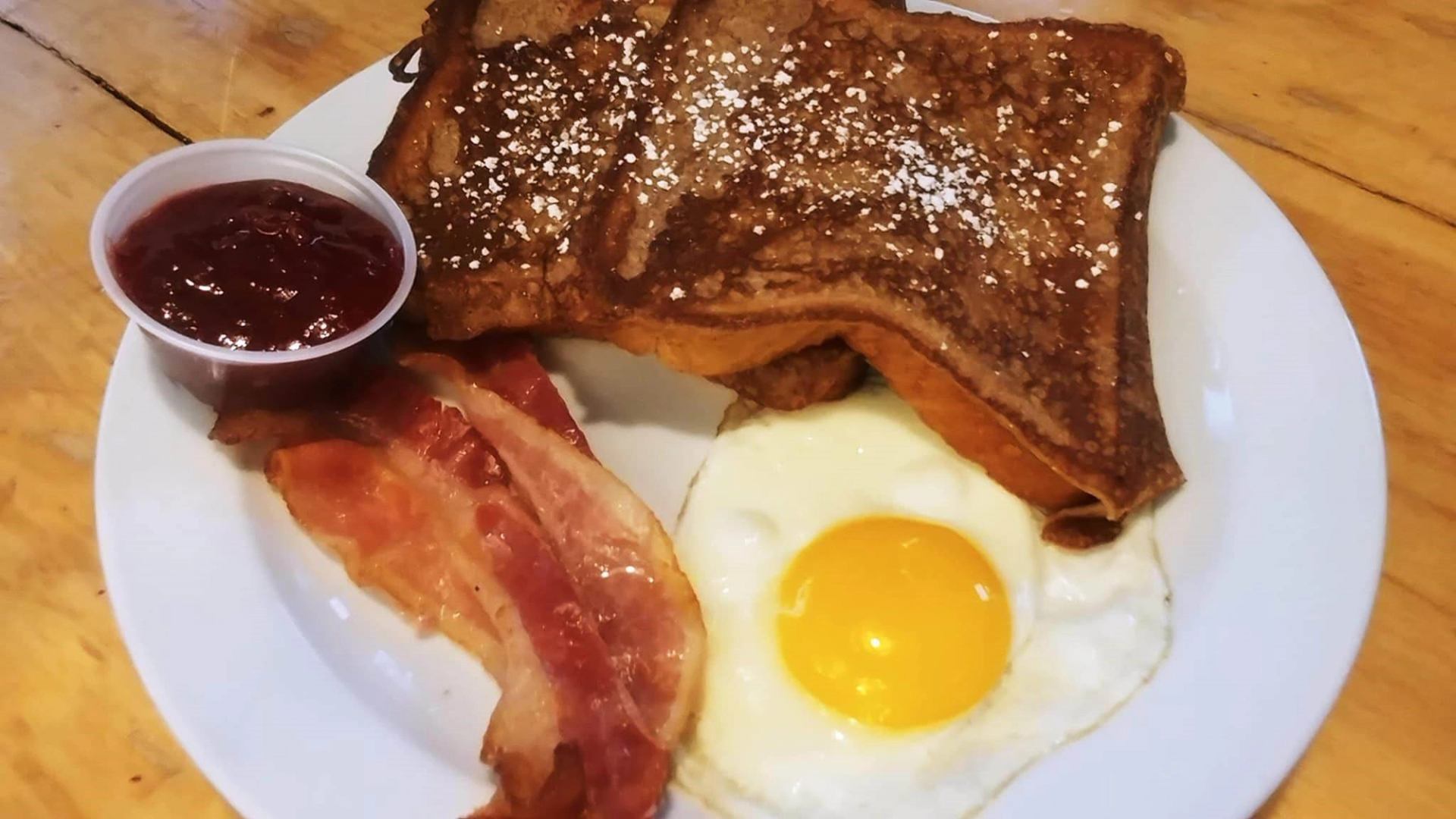 French toast, egg, bacon, and jam served on a white plate at a wooden table.
